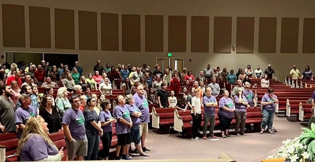 A group of people gathered for a service or event inside a church, some standing and some seated in pews.