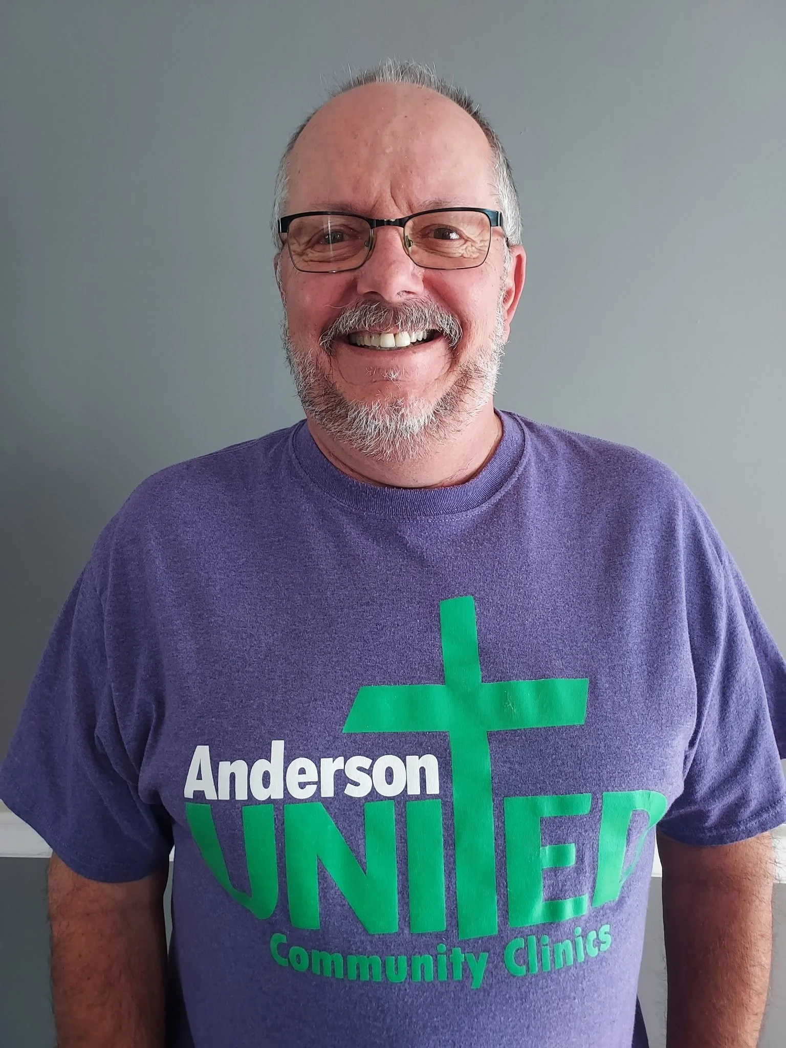 A smiling middle-aged man with glasses and a beard wearing a purple t-shirt with green and white text and cross graphic, standing against a gray background.