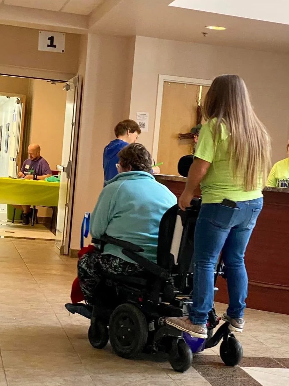 A woman with long hair in a green shirt standing on a mobility scooter, talking to a seated woman in a blue shirt at a reception desk in a healthcare or service facility.