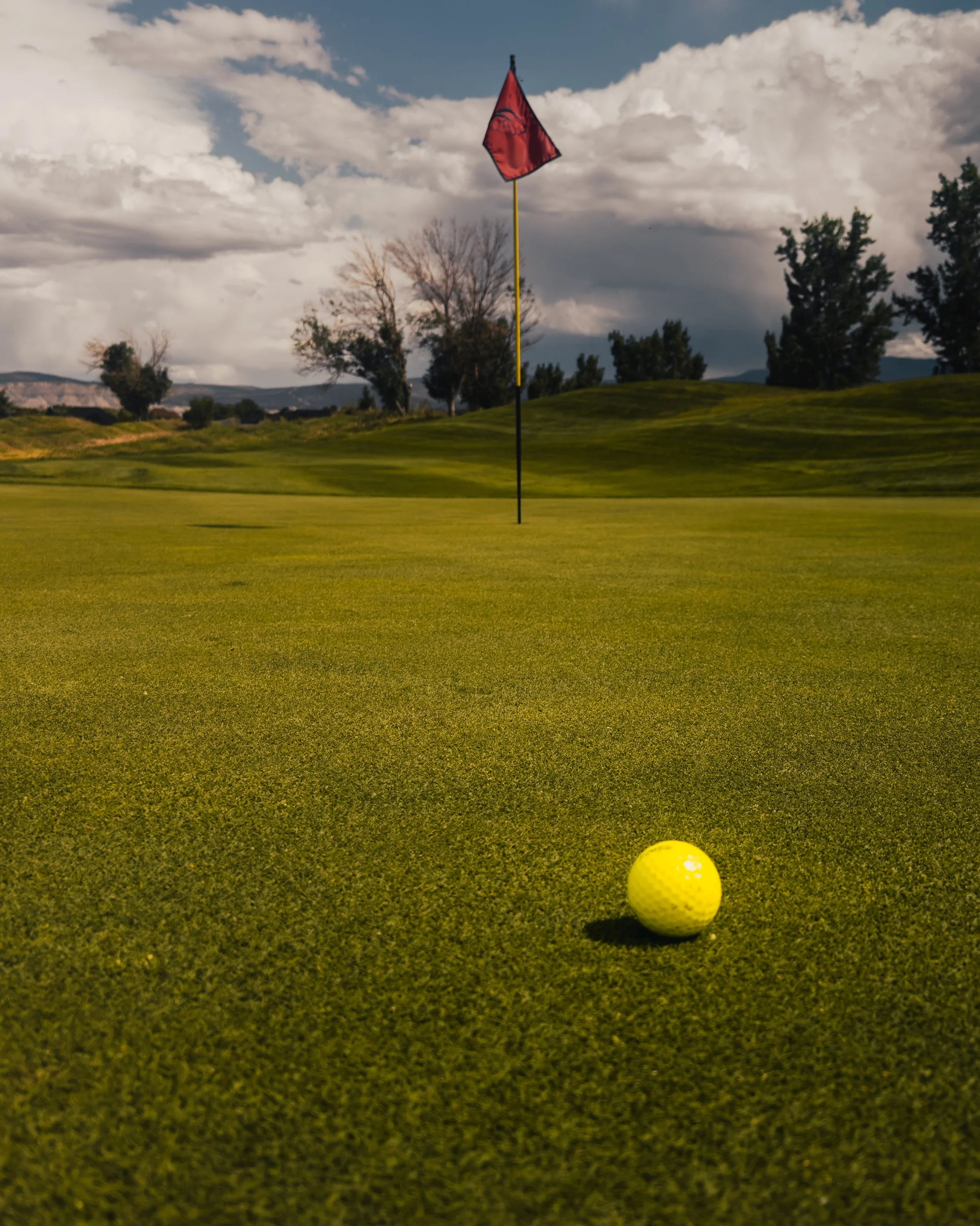 Close-up of a yellow golf ball on a golf green, with a flagstick and red flag in the background, under a cloudy sky surrounded by trees.