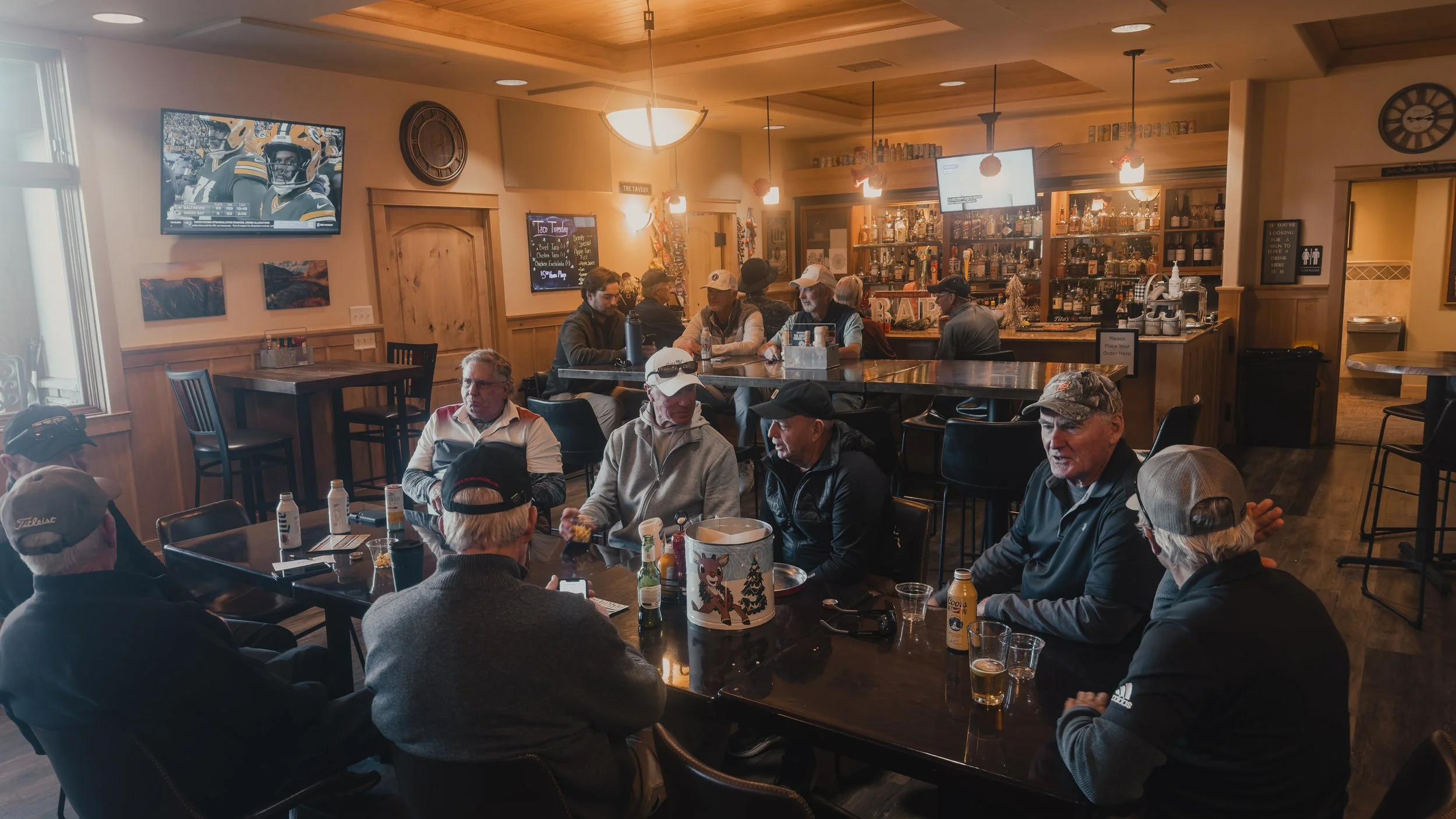 Interior of a cozy bar or restaurant with several groups of older men gathered and talking, some wearing baseball caps. The bar is stocked with bottles, and multiple TVs are on with sports games playing. The room has wooden paneling and warm lighting, creating a welcoming atmosphere.