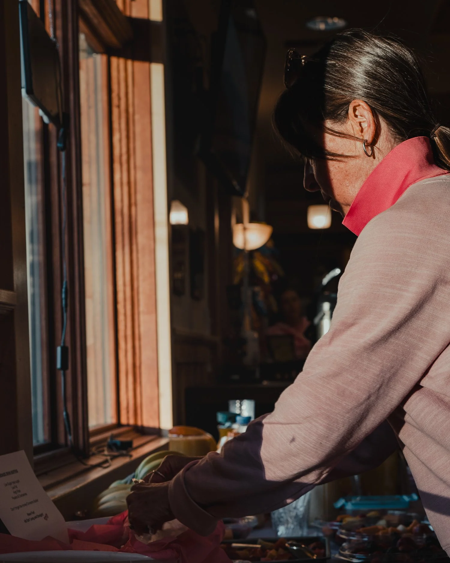 A woman in a pink shirt with dark hair and headphones is in a dimly lit room near a window, handling items on a table.