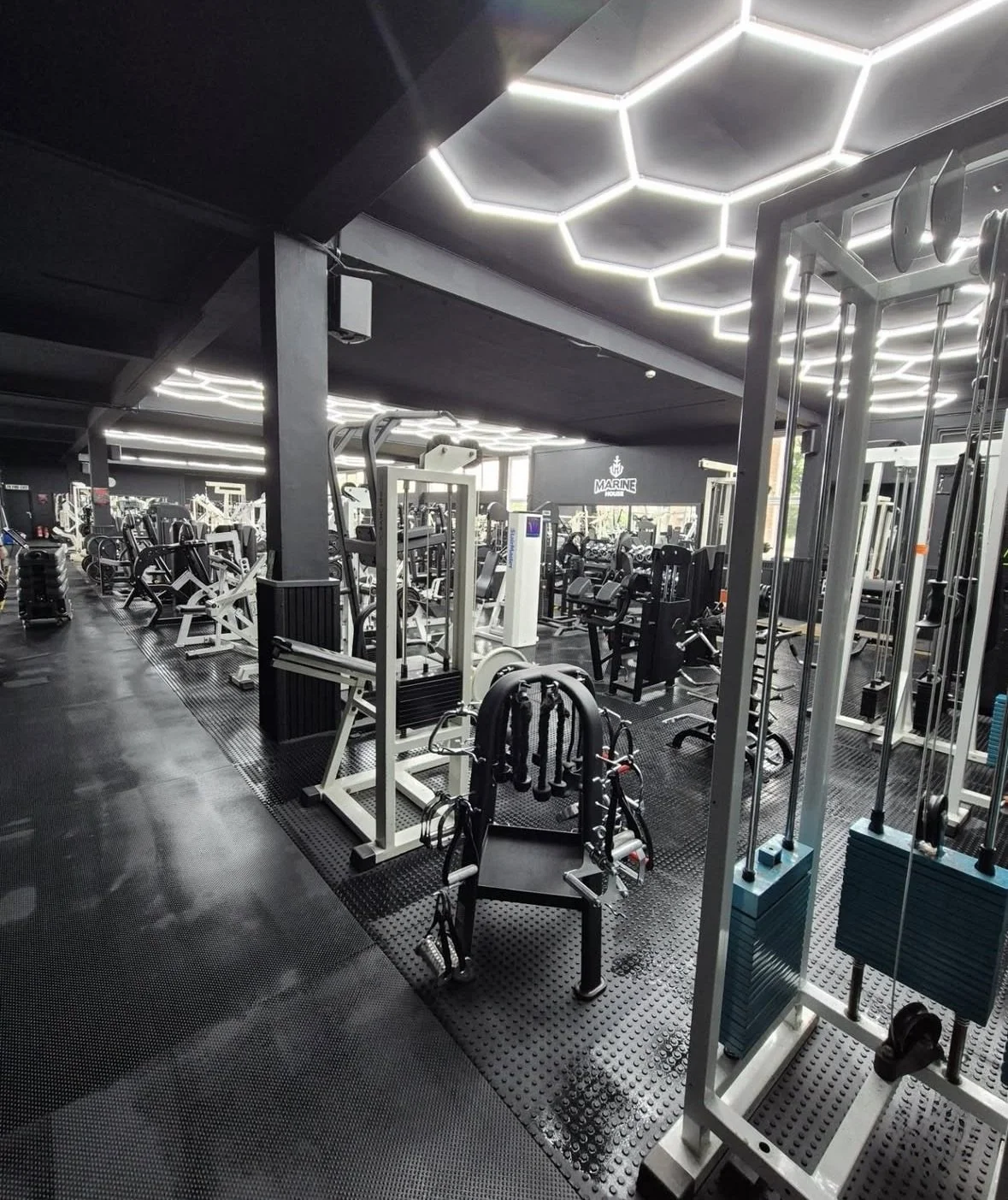 Interior of a modern gym with various workout equipment, black walls, and hexagon-shaped ceiling lights.