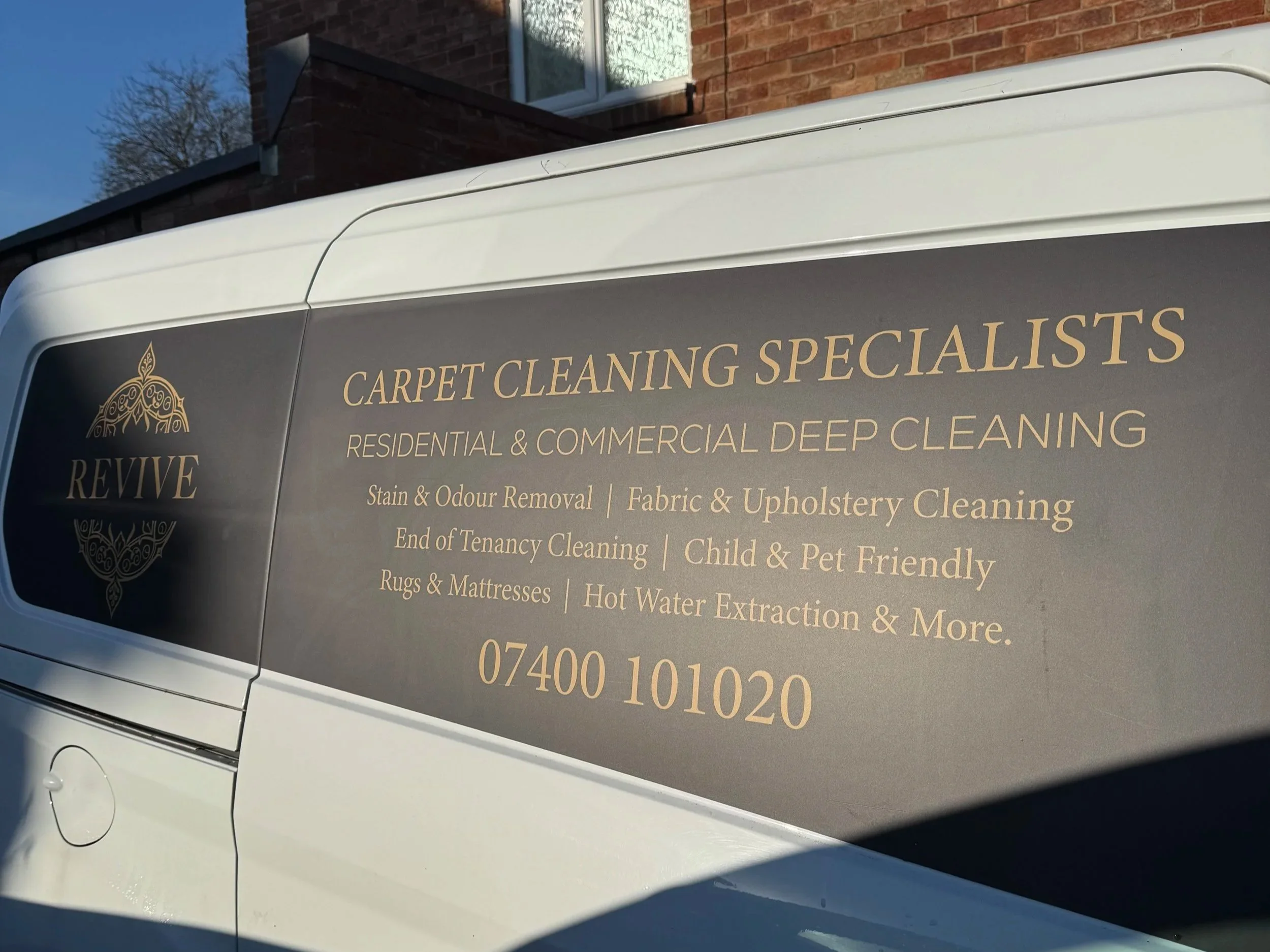 Side of a white van with a gray sign advertising Revive carpet cleaning services, including residential and commercial deep cleaning, stain and odor removal, fabric and upholstery cleaning, end of tenancy cleaning, child and pet friendly service, rugs and mattresses, hot water extraction, and more. Contact number 07400 101020.