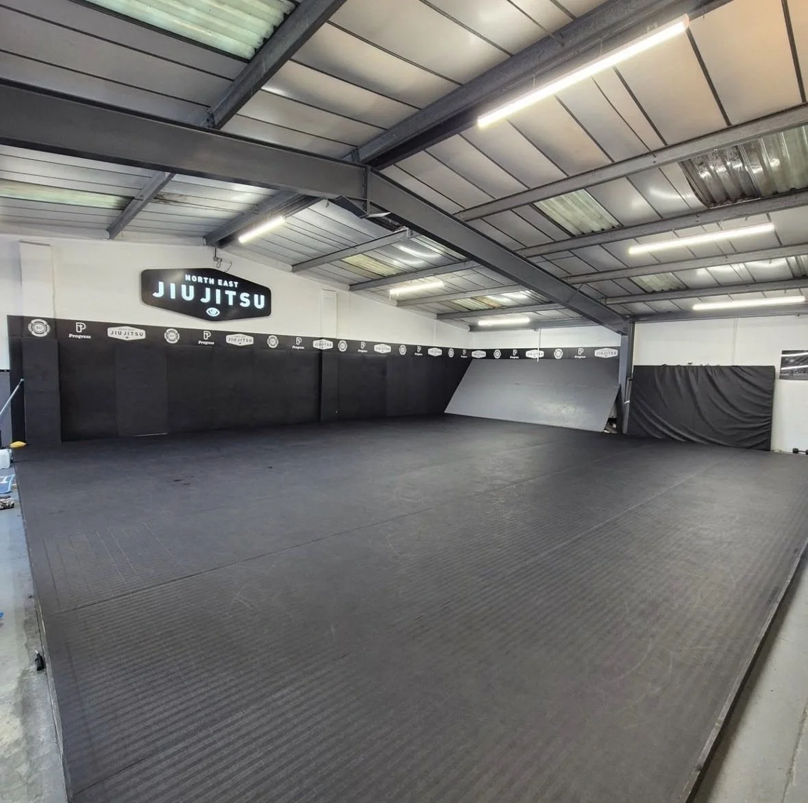 An empty martial arts training area with black padded flooring, black walls with white branding, and a sign that reads 'North East Jiu Jitsu'. The ceiling has metal beams and fluorescent lights.