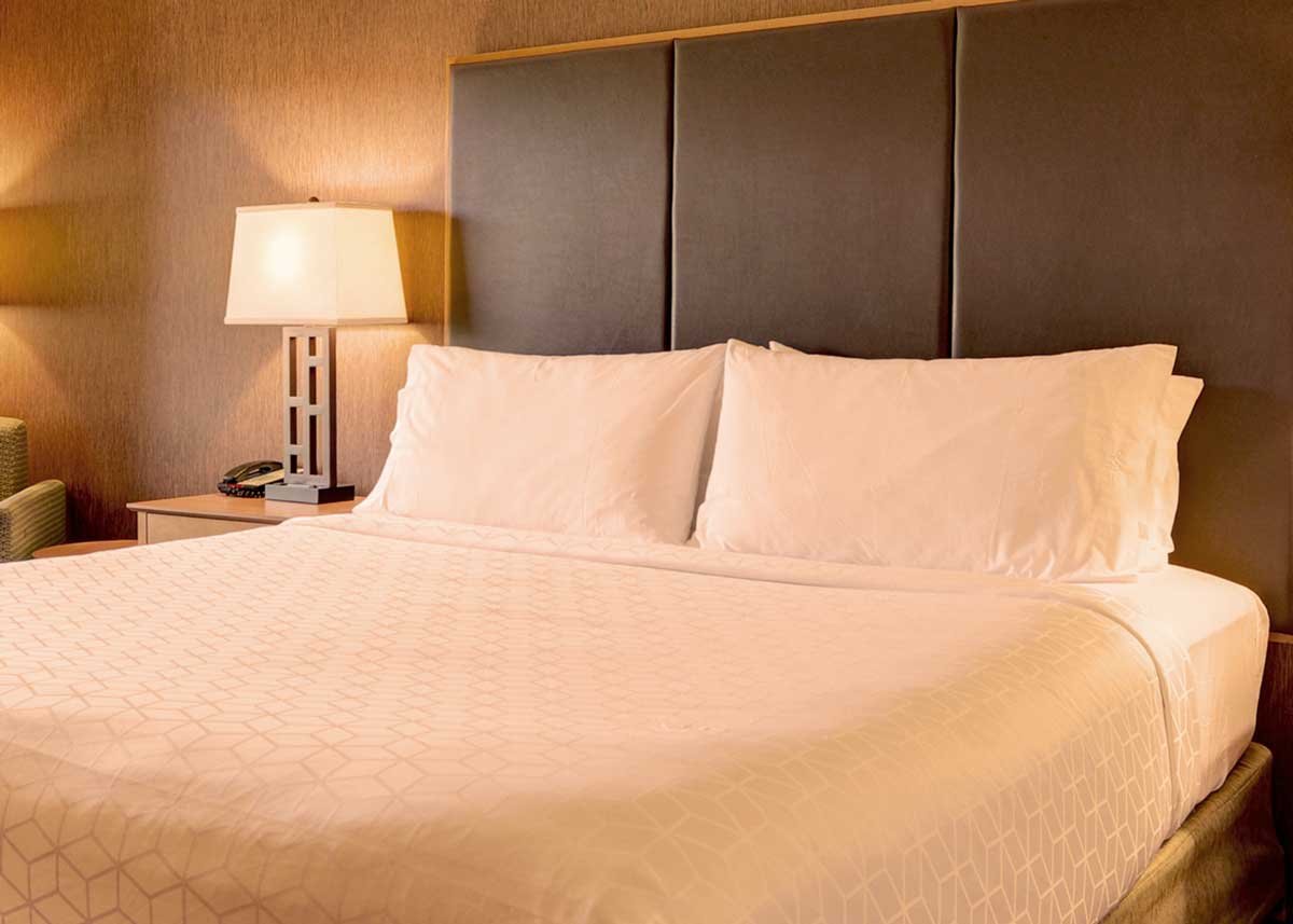 A neatly made hotel bed with an upholstered headboard, nightstand and textured wallpaper