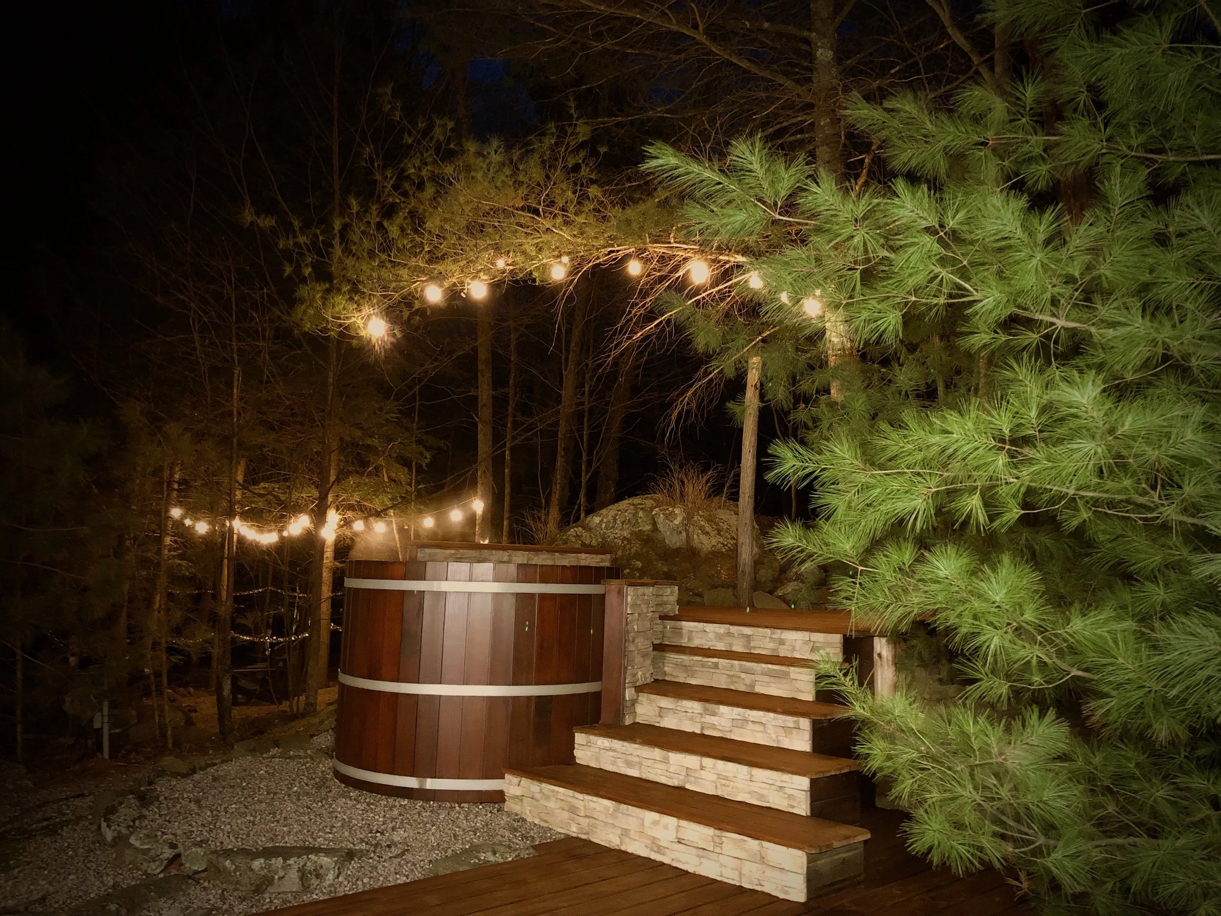 Outdoor hot tub near stone steps and wooden deck, illuminated by string lights at night, surrounded by trees with green foliage.