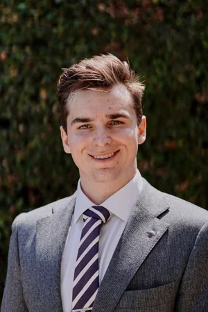 A young man in a gray suit, white shirt, and striped tie smiling outdoors with trees in the background.