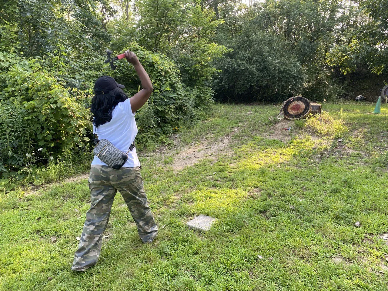 A girl wearing camouflage pants, a white shirt, black cap, and a crossbody bag is hitting a target with an axe in an outdoor wooded area.