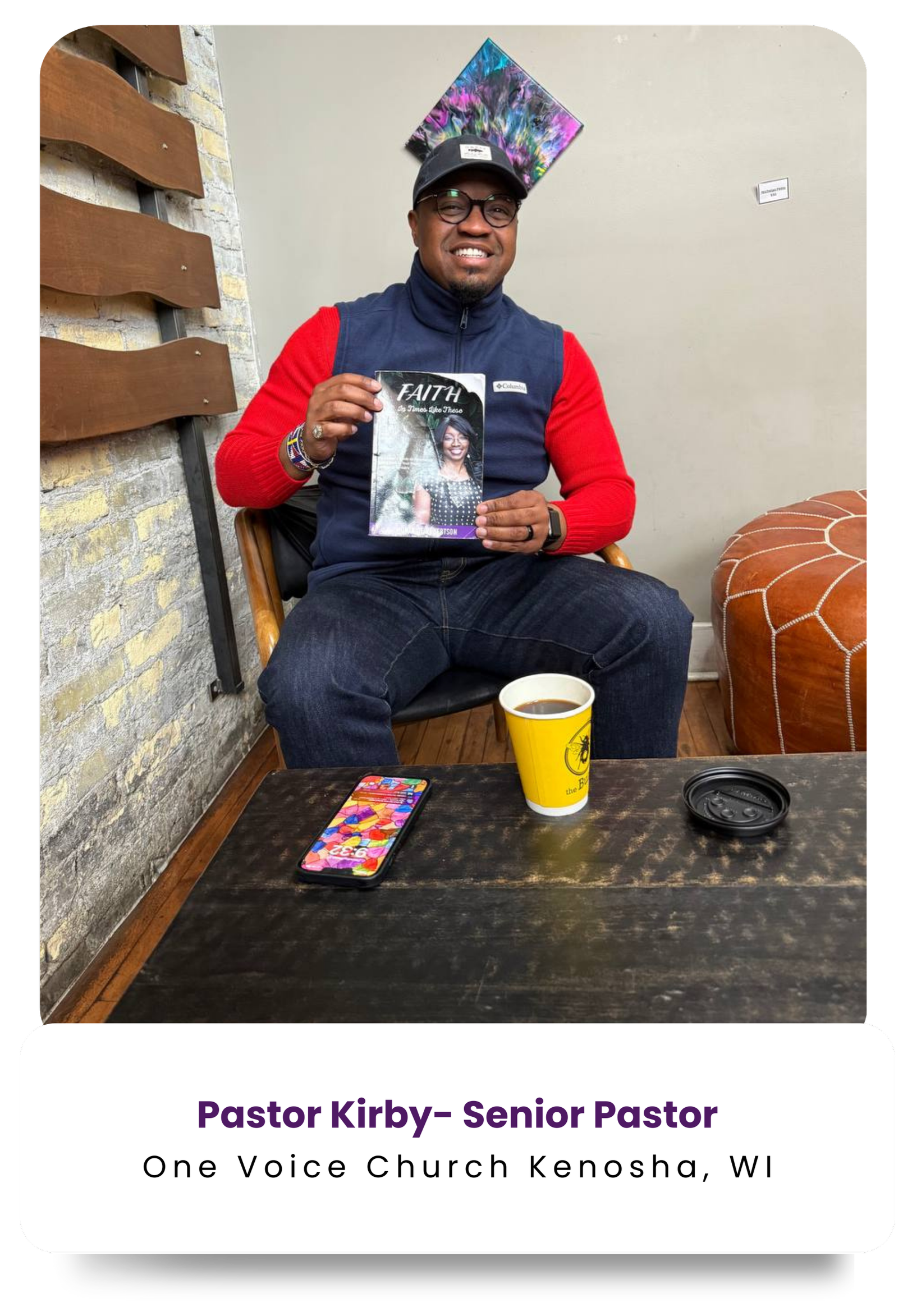 A man wearing glasses, a black cap, a navy vest with red sleeves, and jeans, sitting on a chair. He is smiling and holding a magazine titled 'FAITH'. In front of him on a dark wooden table are a smartphone with a colorful case, a cup of coffee in a yellow cup, and a black lid. The background features a brick wall, a colorful abstract artwork on the wall, and a leather ottoman.