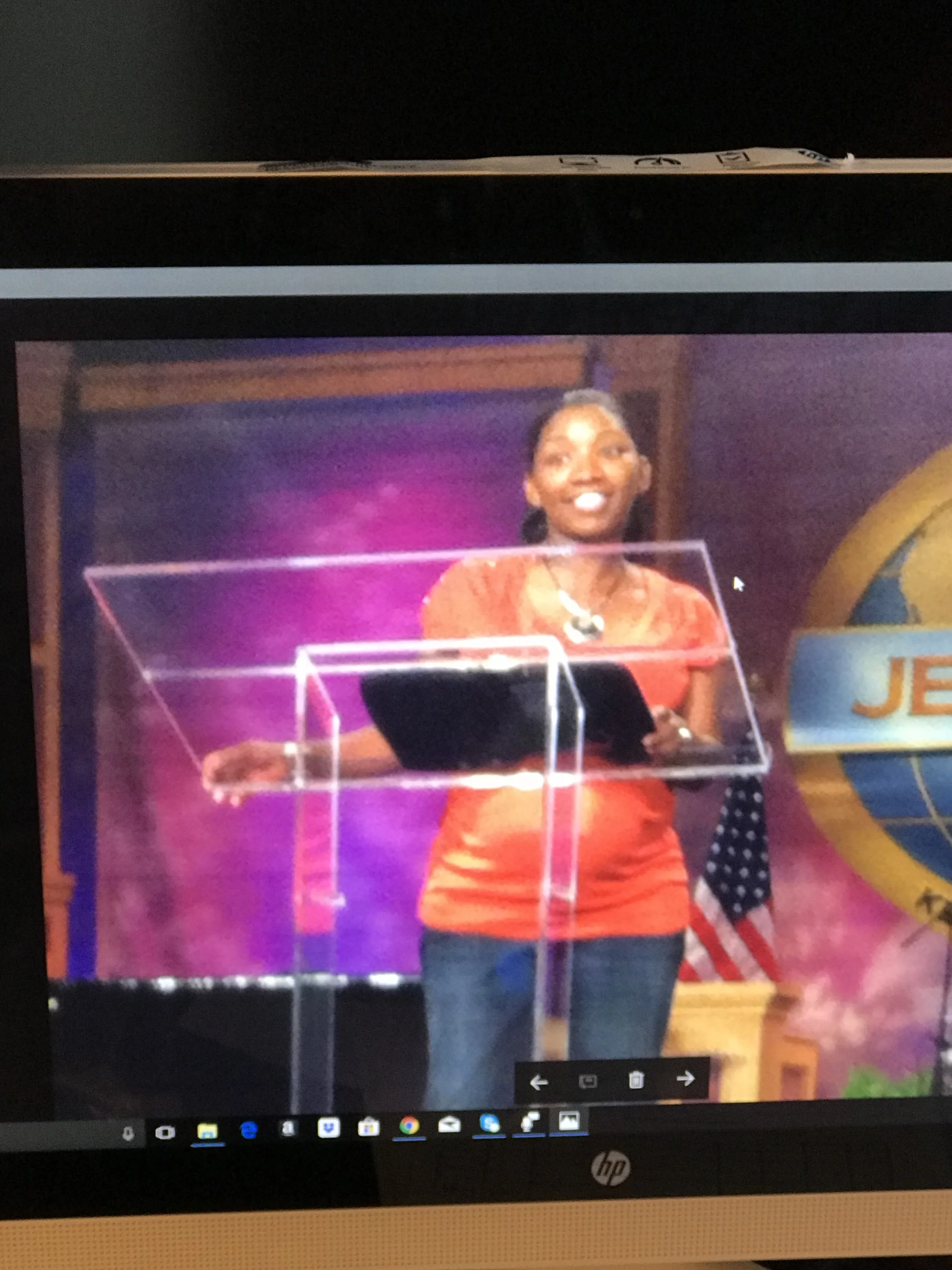 A woman standing on a stage near a podium, with the logo of Jehovah's Witnesses in the background. She is smiling and holding a tablet, wearing a red top and blue jeans.