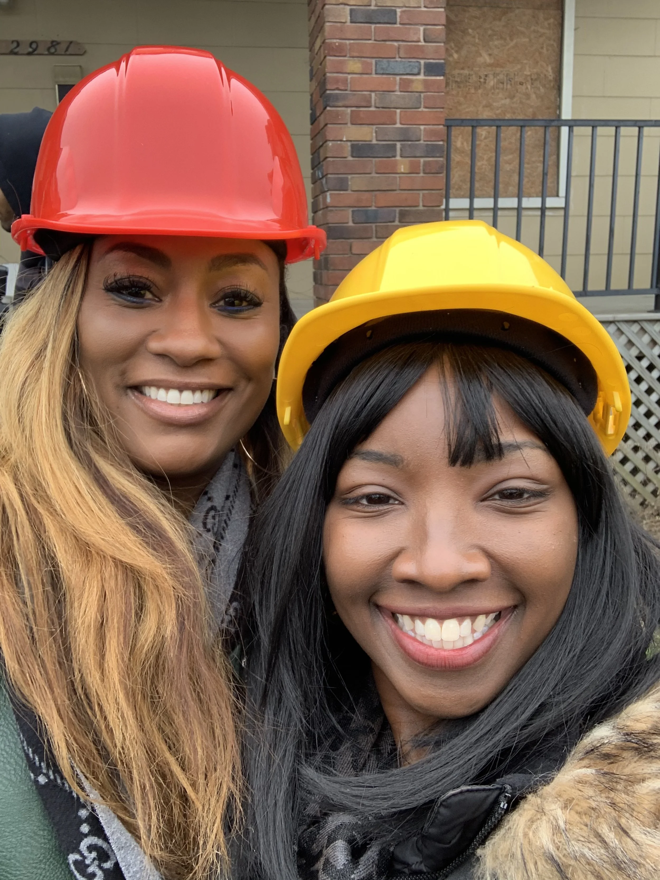 Two women wearing yellow and red construction helmets, smiling, outdoors in front of a brick building.