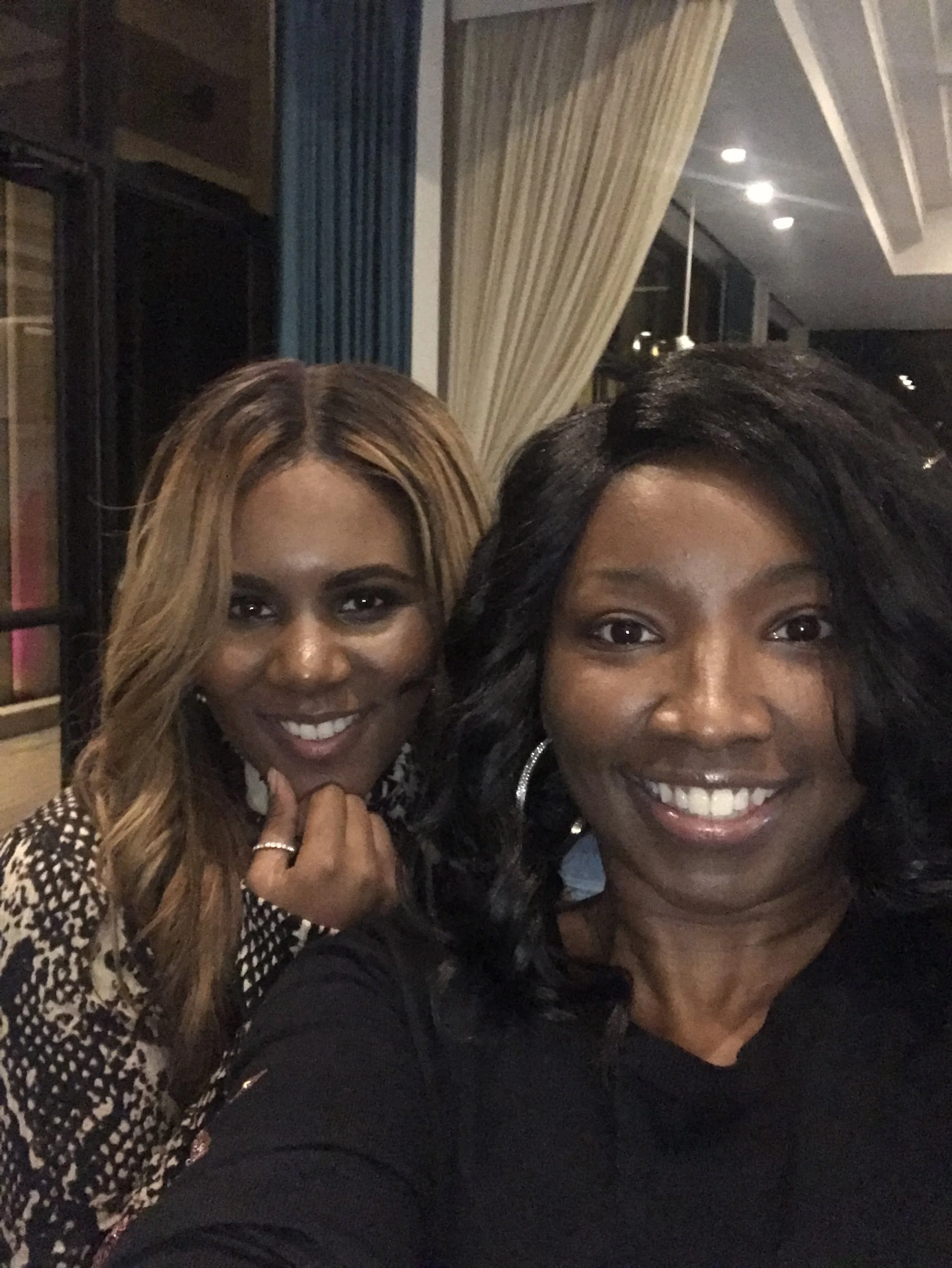 Two women smiling for a selfie indoors with curtains and modern lighting in the background.