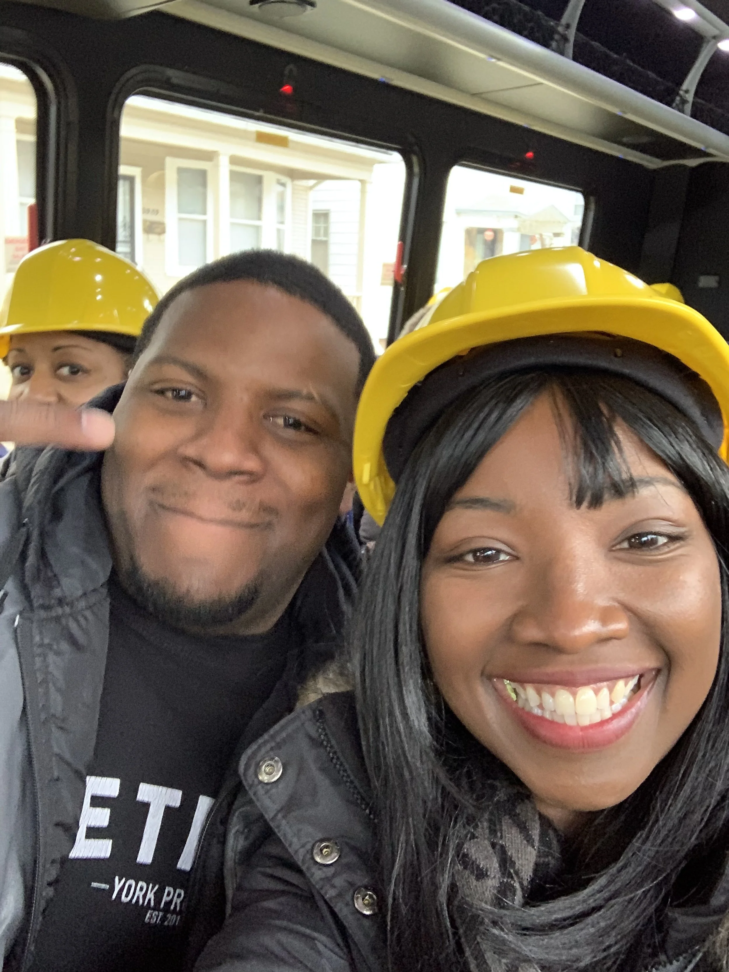 Smiling people, two women and a man, wearing yellow safety helmets, inside a bus or trolley, with a background of houses visible through the windows.