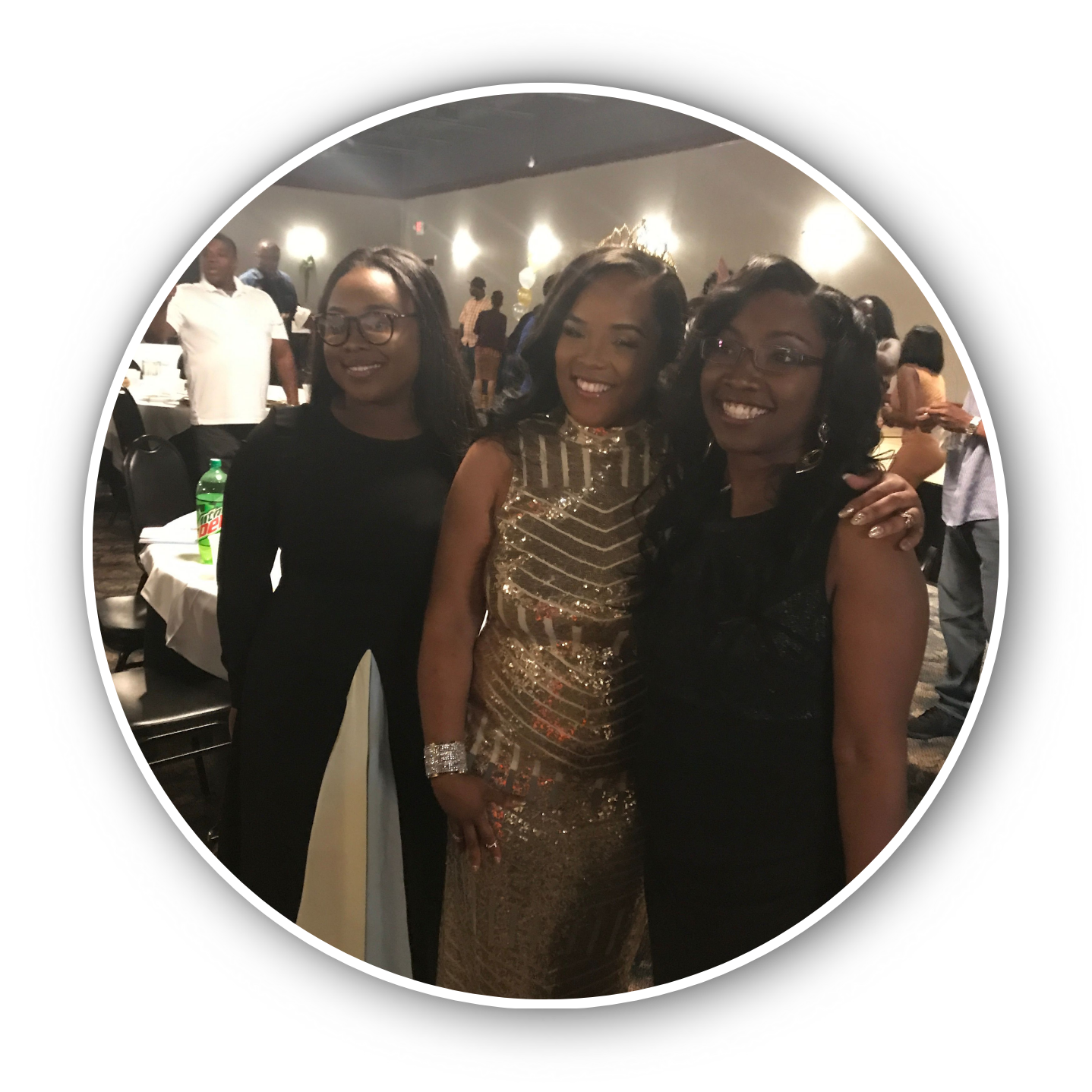 Three women smiling at an indoor event, one in a black dress, one in a gold dress, and one in a black lace dress, with people and tables visible in the background.