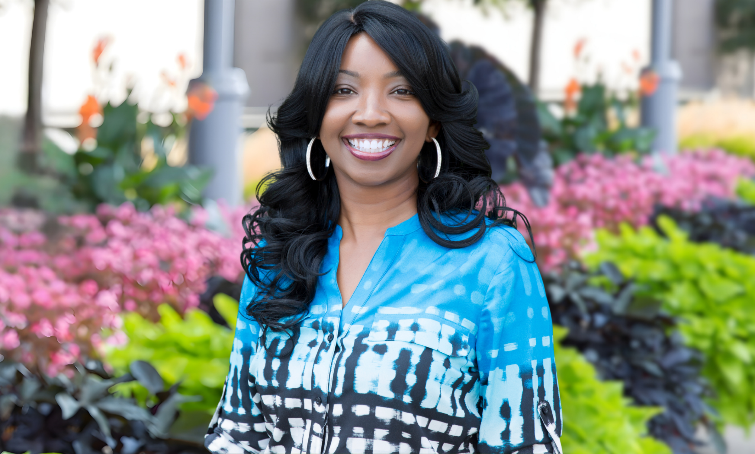A woman with long black hair and hoop earrings smiling outdoors with colorful flowers and greenery in the background.