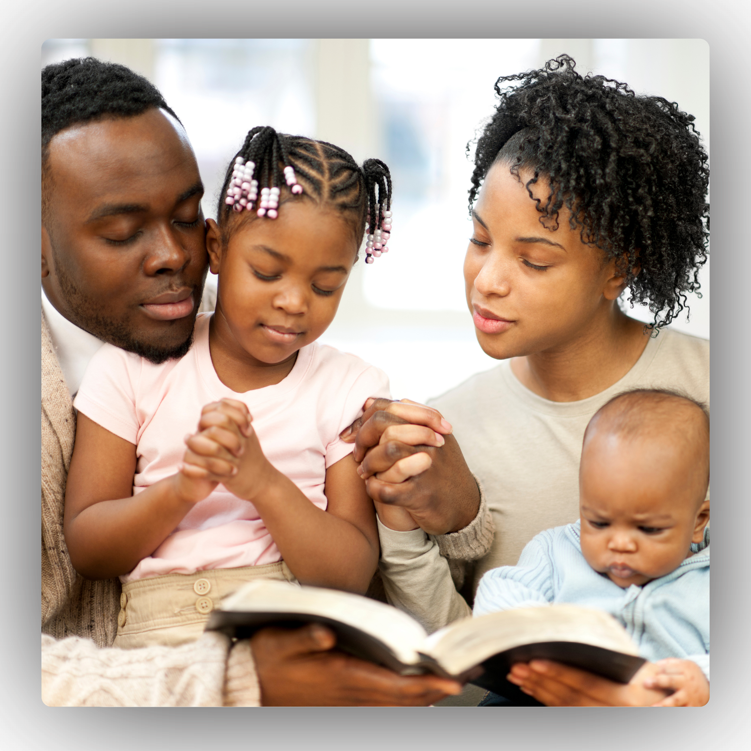 Family of four reading a book together, with two children and two adults, all engaged and praying.