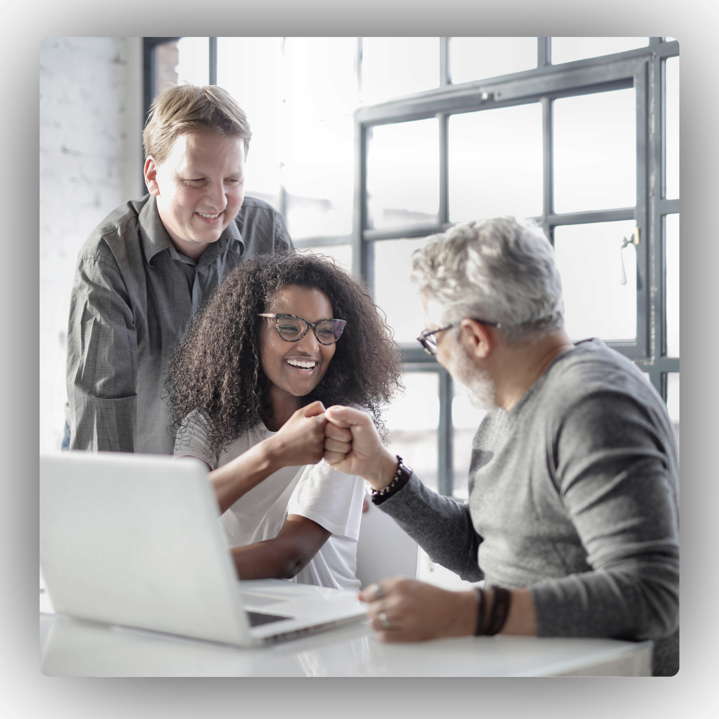 Four colleagues, two men and two women, are in an office with large windows. They are engaged in a friendly arm-wrestling match at a desk, smiling and cheerfully interacting.