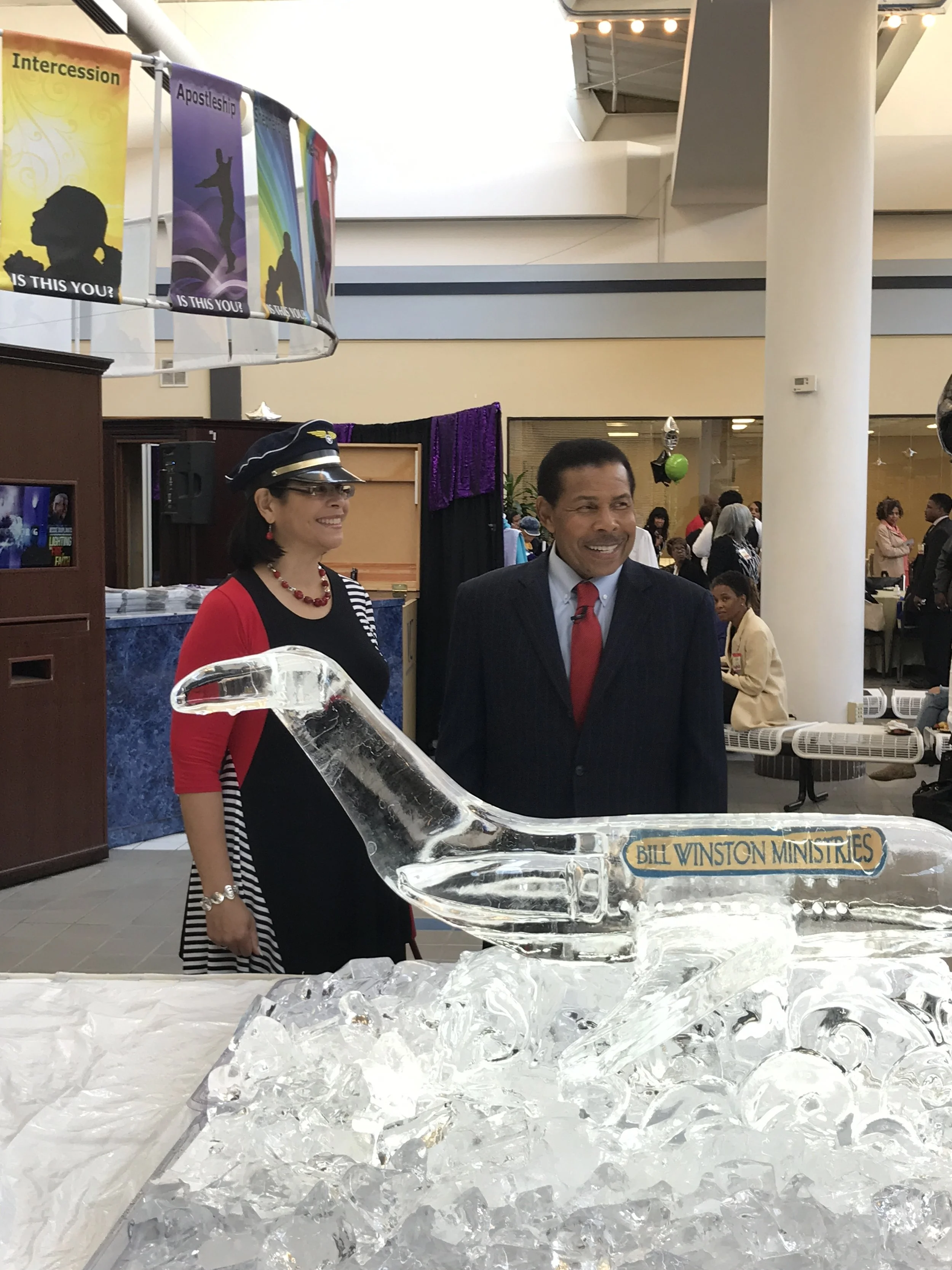 People standing near a table with an ice sculpture of a dinosaur labeled "Bill Winston Ministries" in an indoor event space.