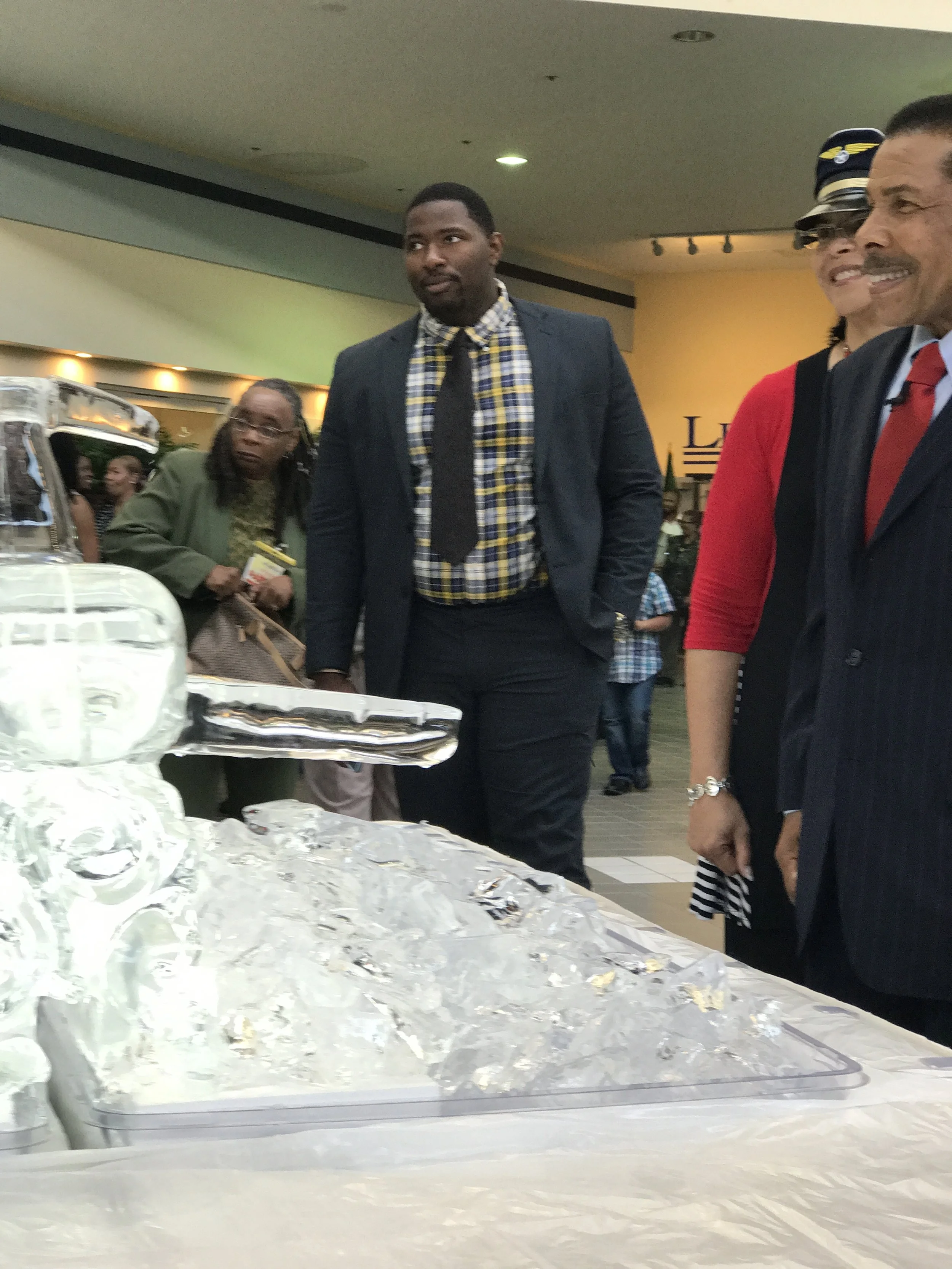 People gathered around a table with a large tray of ice, possibly containing a cake or desserts, in an indoor setting.