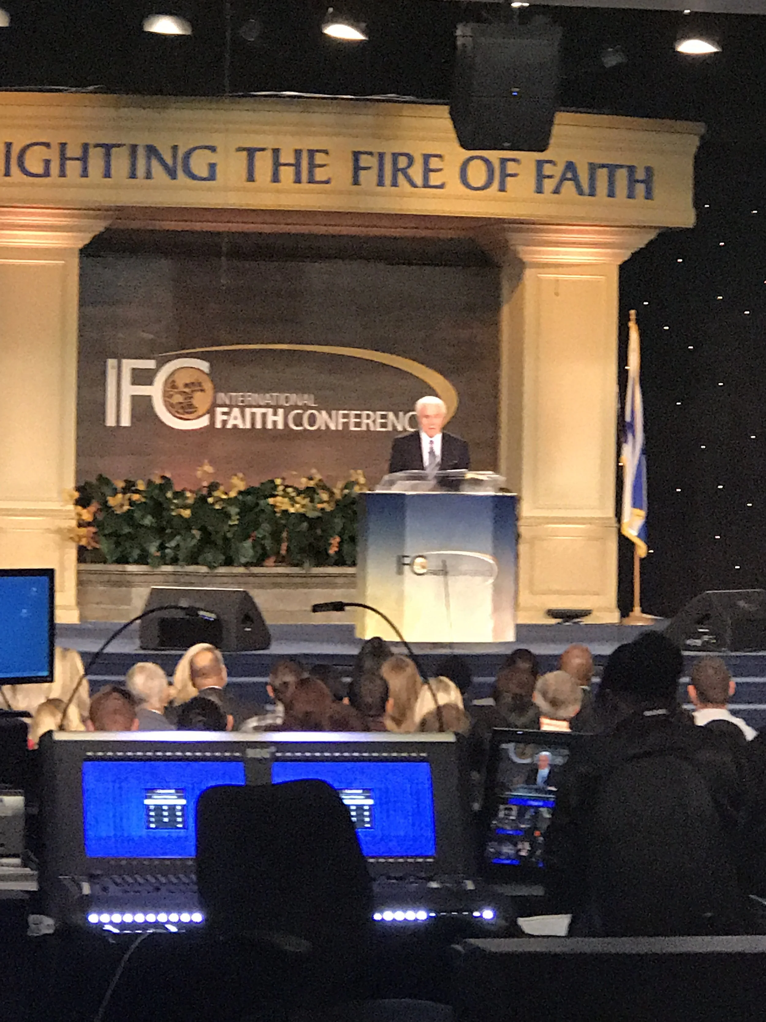 A speaker on stage at the International Faith Conference, standing behind a podium with the conference logo. The stage displays a banner that reads 'FIGHTING THE FIRE OF FAITH' and features plants with yellow flowers. The audience is visible in the f