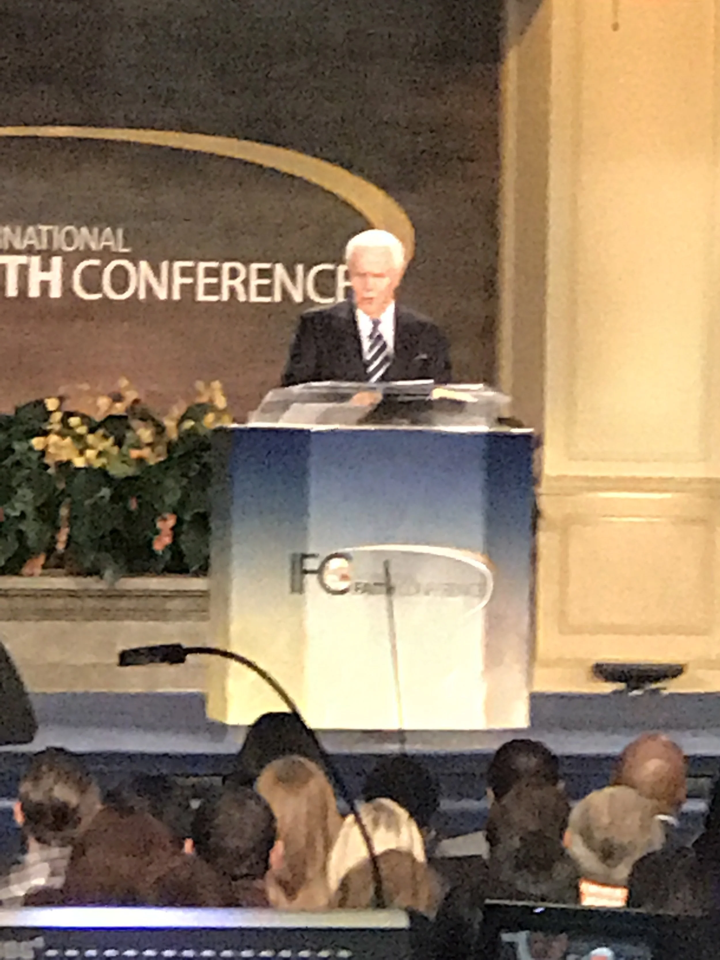 A man on stage giving a speech at the International Speech Conference, behind a podium with the conference logo, and an audience seated watching.