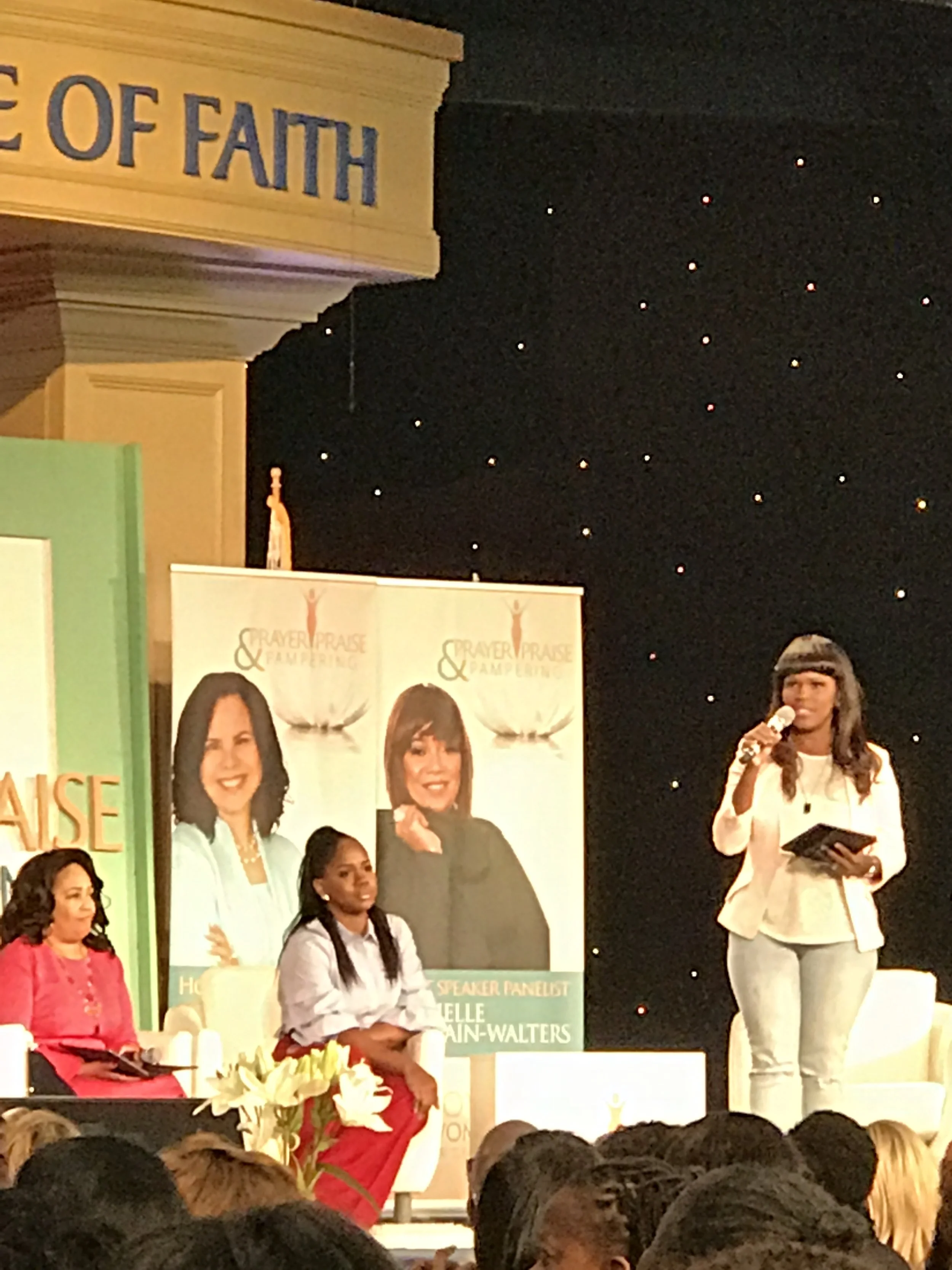 A woman holding a microphone and reading from a tablet on stage at a conference or seminar. Behind her are two large posters with images of women and text, and a seated woman in a red outfit. The background is a dark starry curtain, and part of a sig