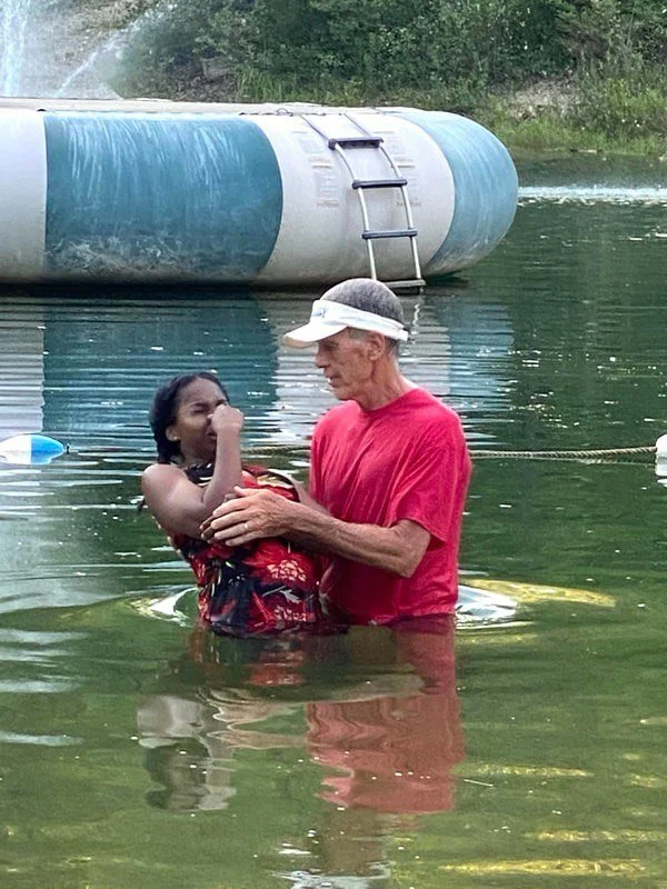 An elderly man and a young woman are standing in a body of water, with the man holding the woman as she appears to be crying or upset. A large floating water tank or buoy and a ladder are visible in the background.