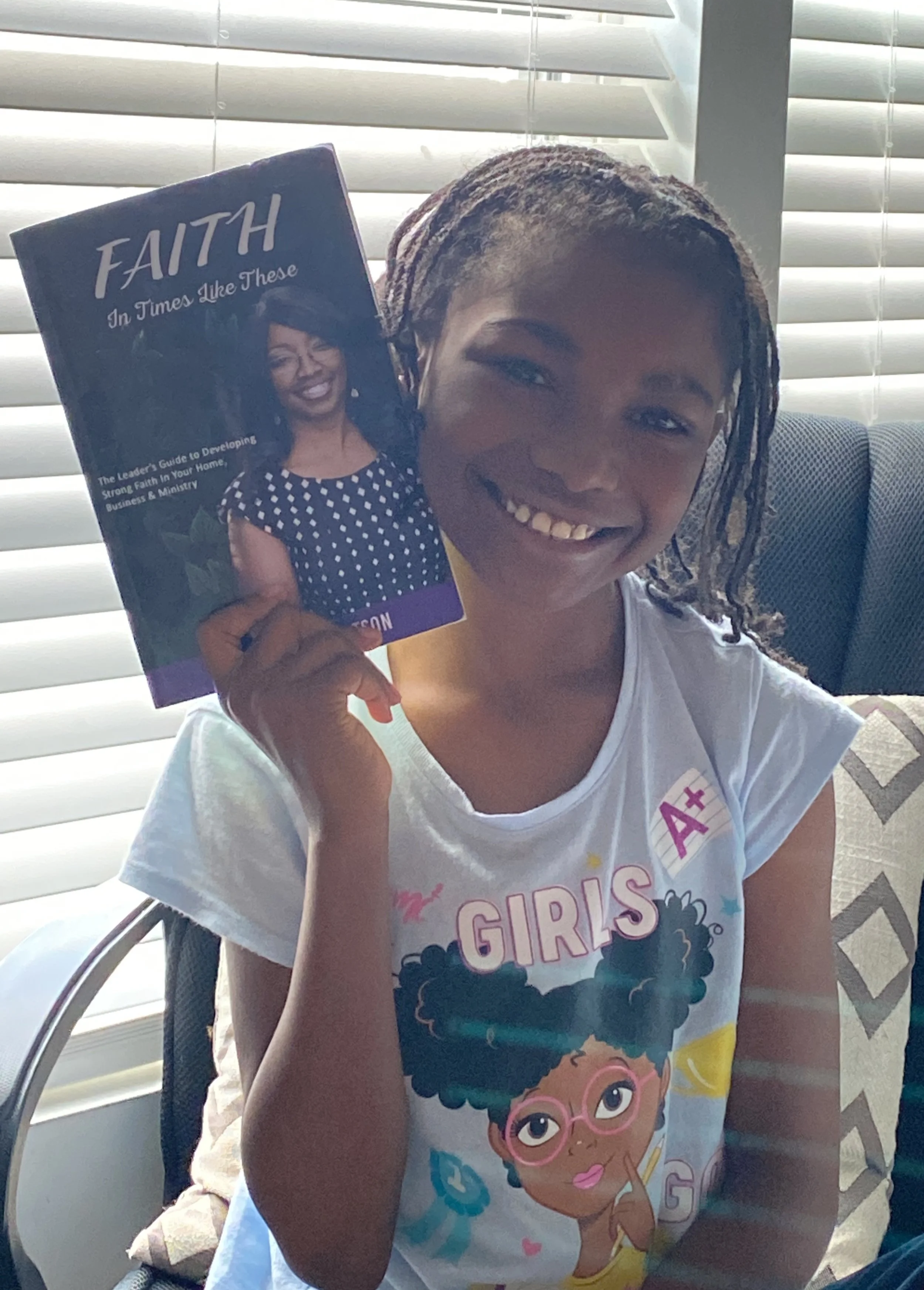 Young girl smiling and sitting on a chair, holding a magazine titled 'FAITH In Times Like These' window blinds and a cushioned armchair in the background.