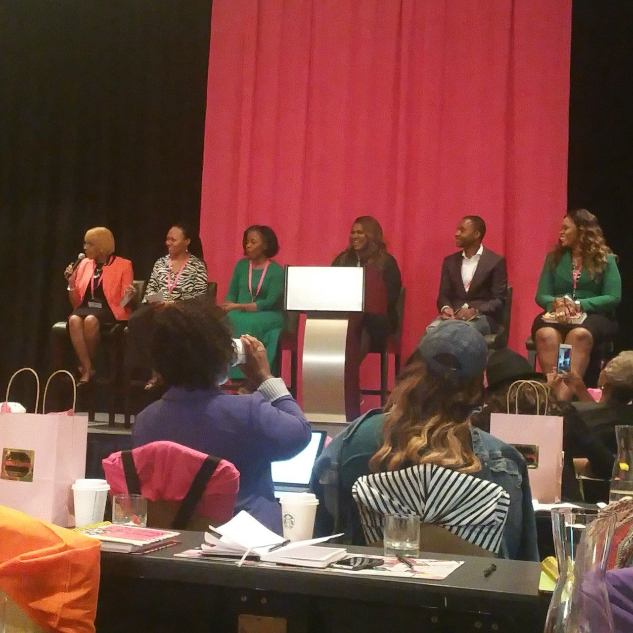 A panel of six diverse women sitting on stage at a conference, with a large pink curtain behind them. Audience members are seated at tables in front, some taking photos or using laptops.