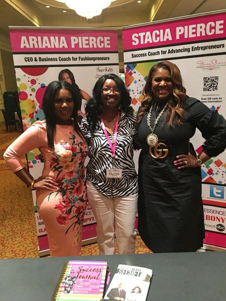 Three women standing in front of two pink banners, smiling at an event. The banners read "Ariana Pierce" and "Stacia Pierce," with titles related to coaching and entrepreneurship. A table in front holds a pink journal titled "The Success Journal" and a magazine titled "The BIG LEAP." The women are dressed stylishly, with the woman on the right wearing a black dress with a Gucci belt and a large necklace.