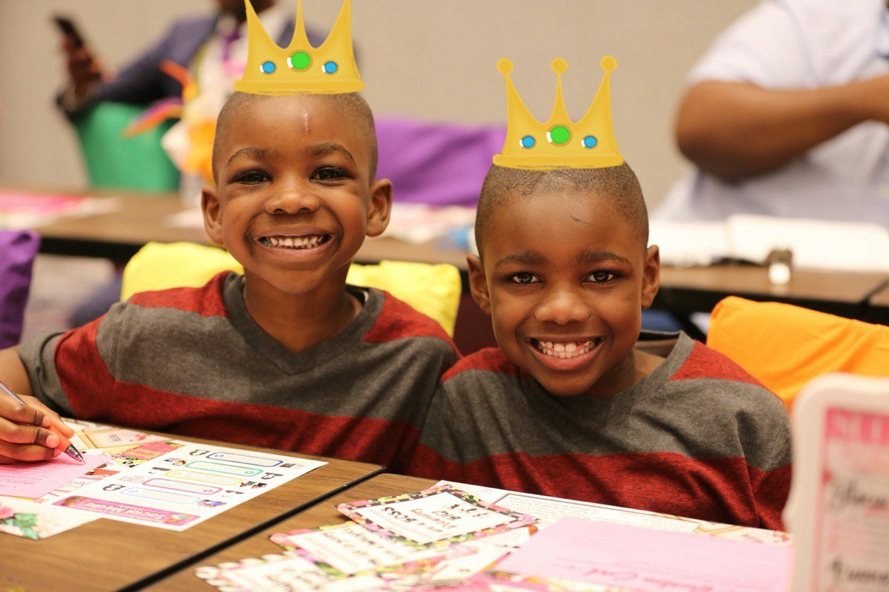 Two children sitting at a table, smiling, wearing paper crowns with colorful jewels, in a classroom or party setting.