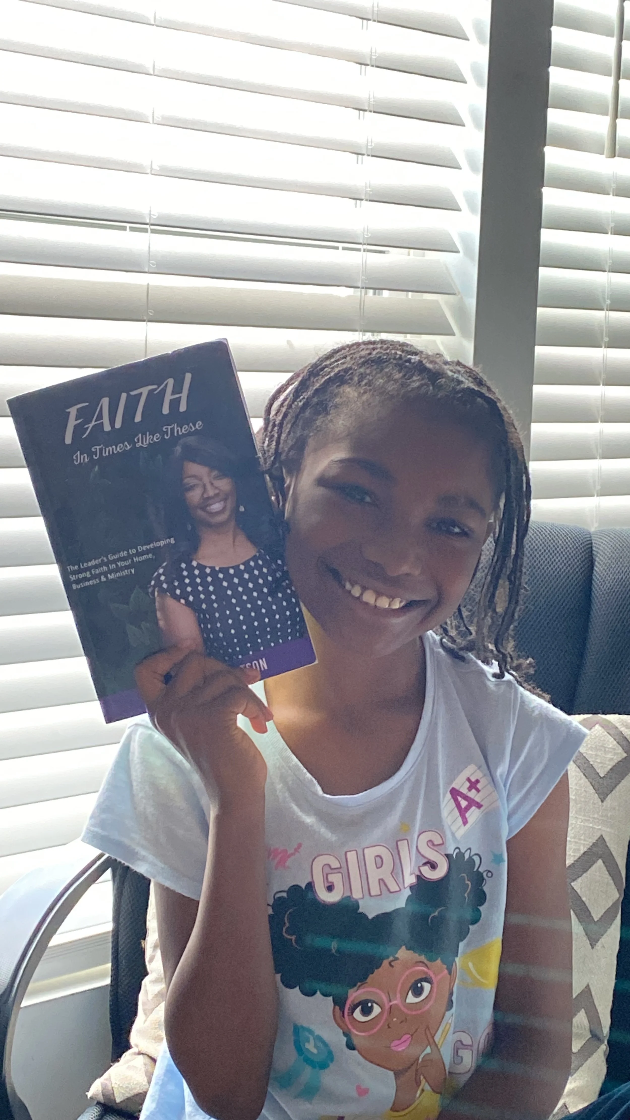 A young girl with braided hair smiling while holding a book titled "FAITH In Times Like These" in front of a window with blinds.