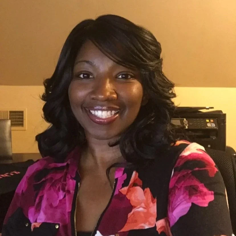 A woman smiling with dark wavy hair wearing a colorful floral top, sitting in an indoor setting with a printer and wall vent in the background.