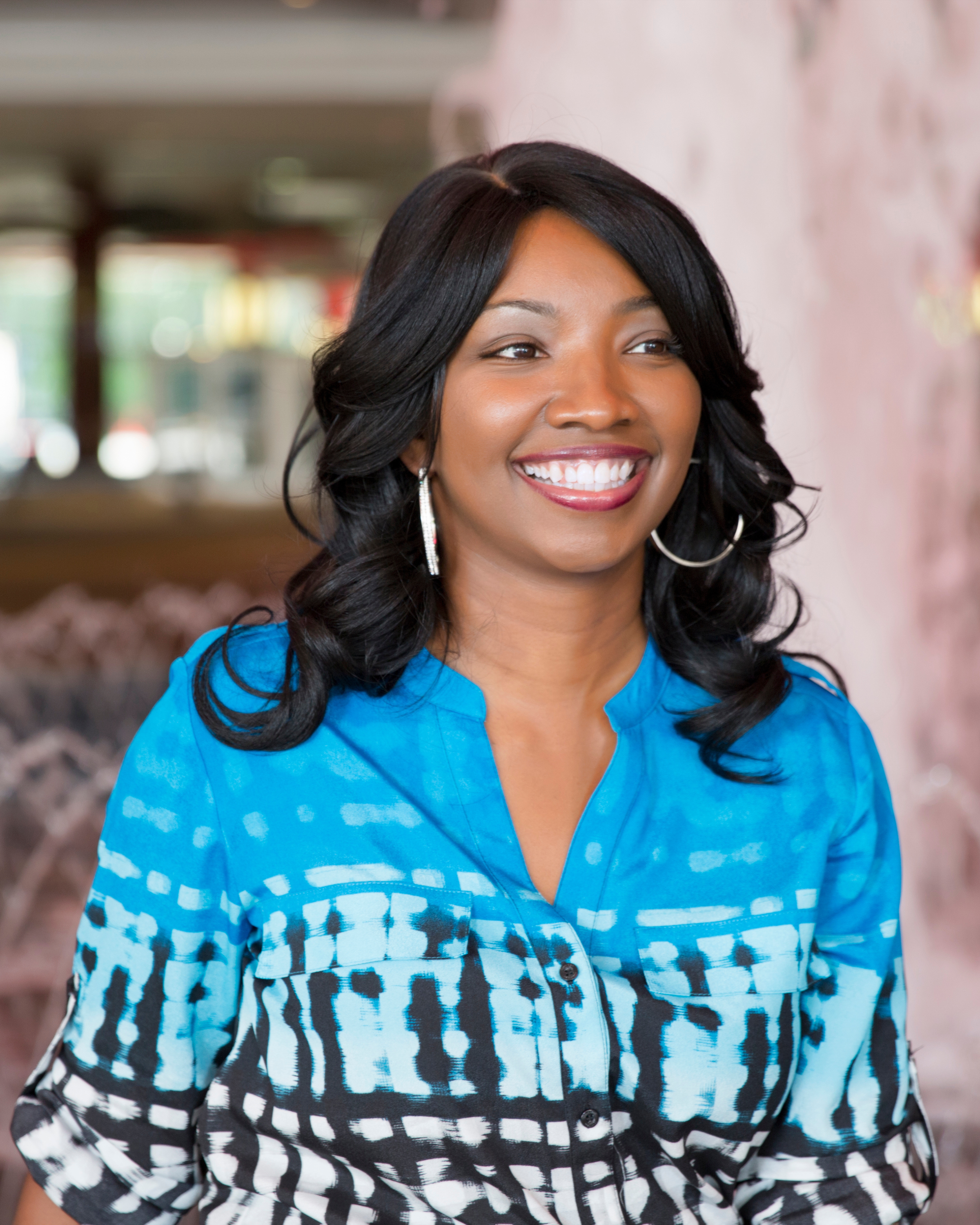 A smiling woman with long black hair, wearing a blue and black patterned shirt and hoop earrings, standing indoors.