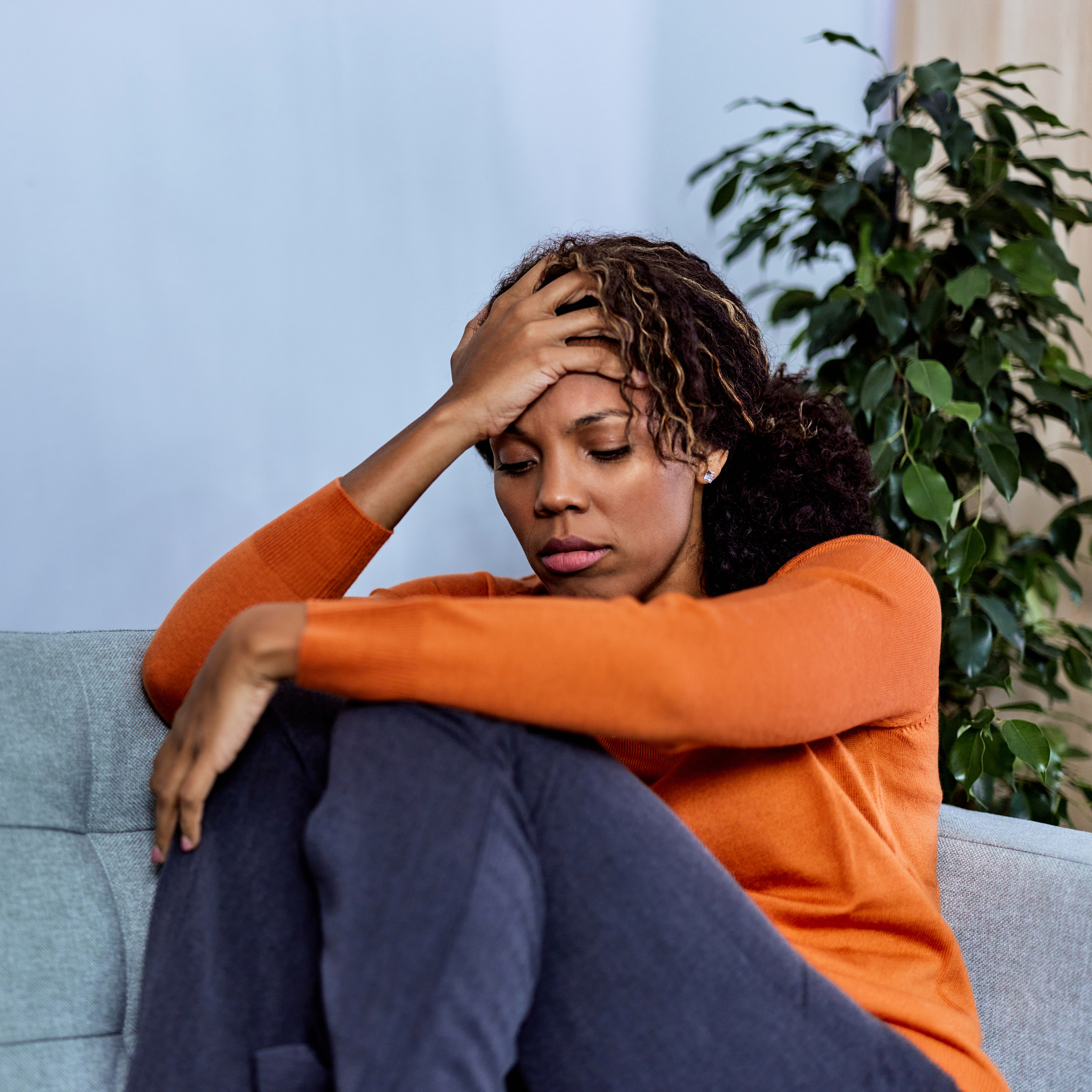 A woman sitting on a couch holding her head with her hand, appearing distressed or worried, with a plant in the background.