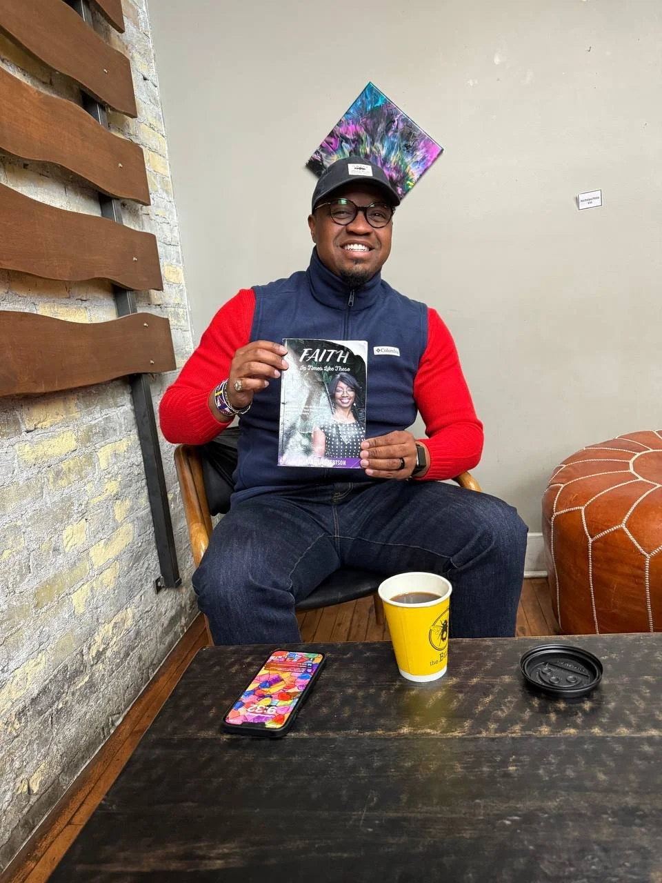 A man sitting at a table in a cafe, smiling and holding a magazine titled 'Faith.' He is wearing glasses, a baseball cap, a red and navy blue jacket, and jeans. On the table, there is a smartphone with a colorful case, a yellow cup of coffee, and a black plastic lid. Behind him, there is a wall with a colorful abstract painting and a decorative leather ottoman.