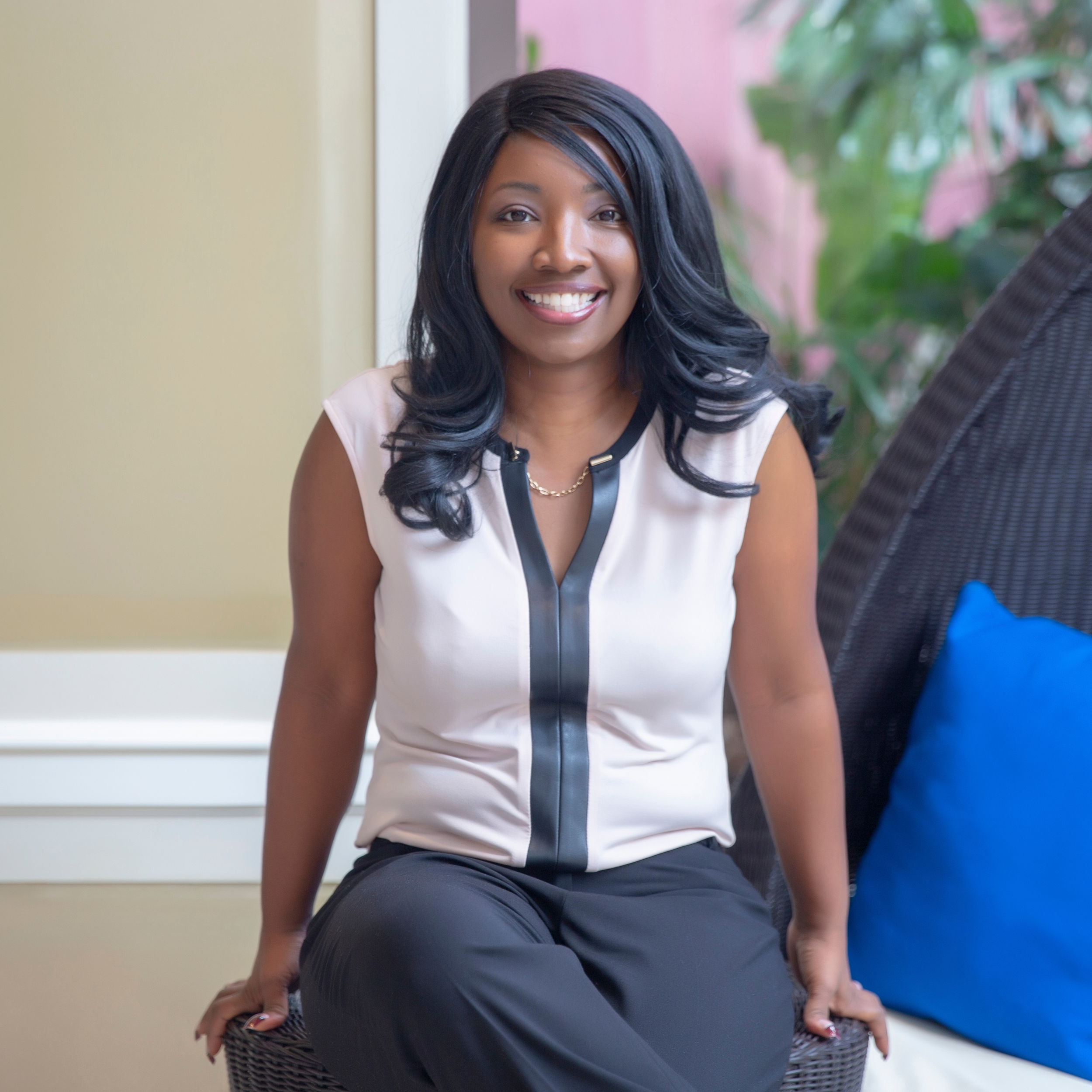 A woman with long black hair, wearing a sleeveless top with black and beige color blocking, sitting on a wicker chair outdoors, smiling at the camera near a window with green plants in the background.