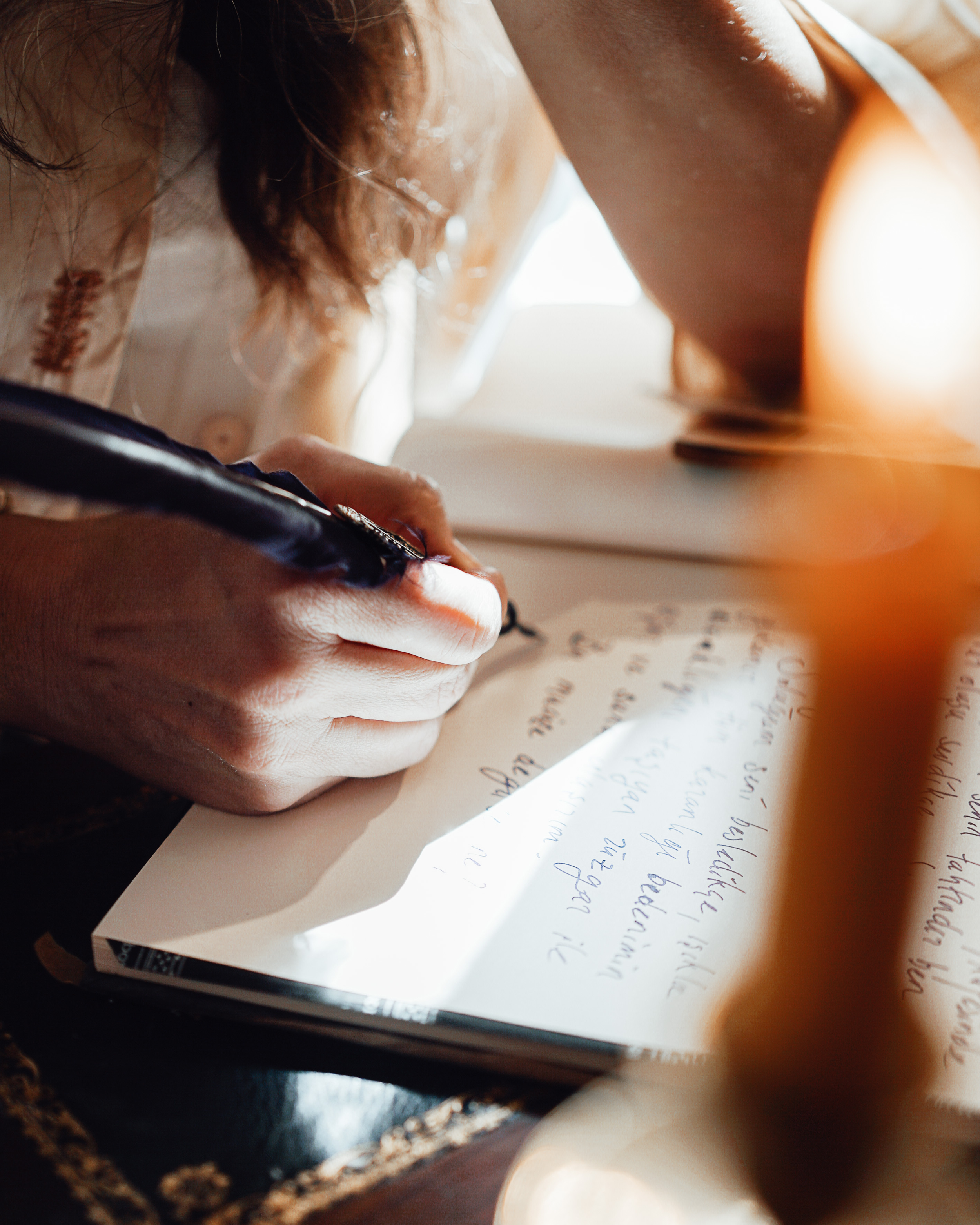 A person writes in a notebook with a black pen while sunlight illuminates the area, capturing part of their hand and face, with fingers visible and a cozy indoor setting.