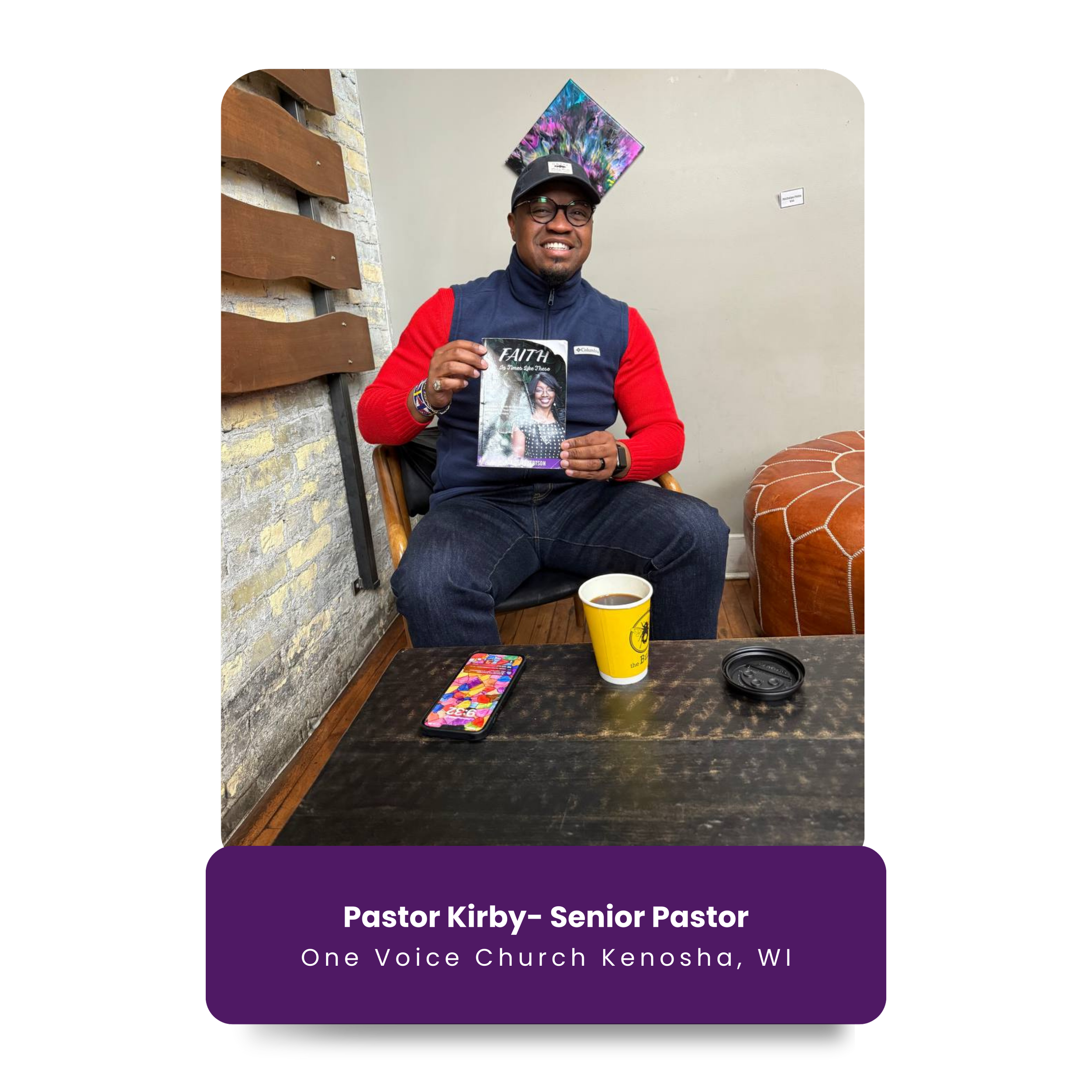 Pastor Kirby, a senior pastor at One Voice Church in Kenosha, WI, sitting in a chair in an indoor space. He is smiling and holding a book titled 'Faith'. There is a cup of coffee and a smartphone on the table in front of him. The background includes 