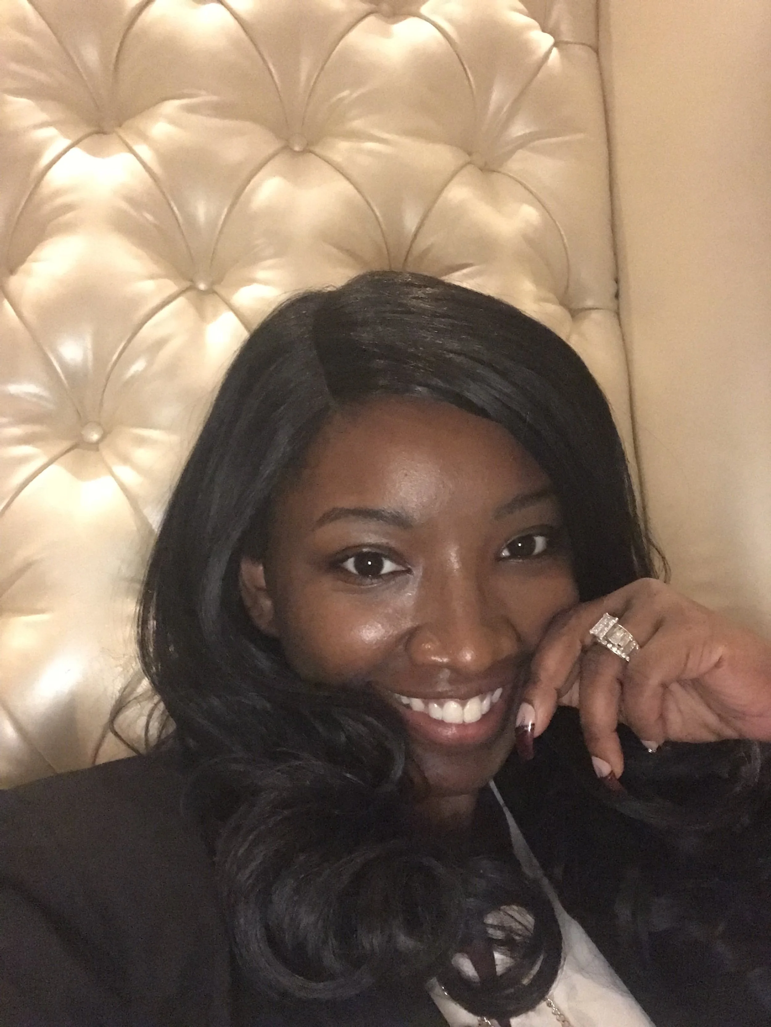 Smiling woman with dark hair, wearing a ring, lying on a cream-colored tufted headboard.