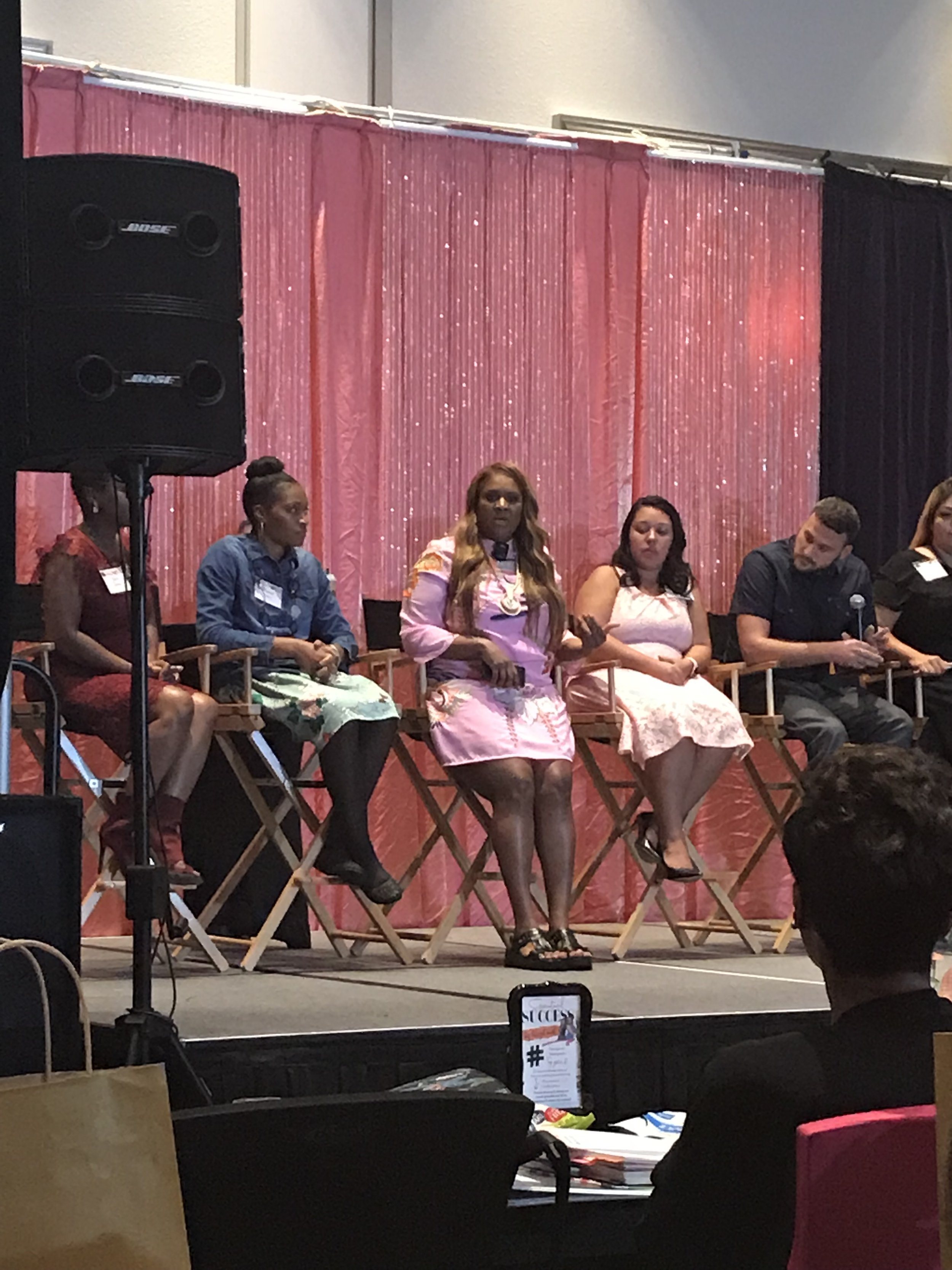 Panel of five people sitting on chairs on stage, with a pink and black backdrop, at a conference or panel discussion.