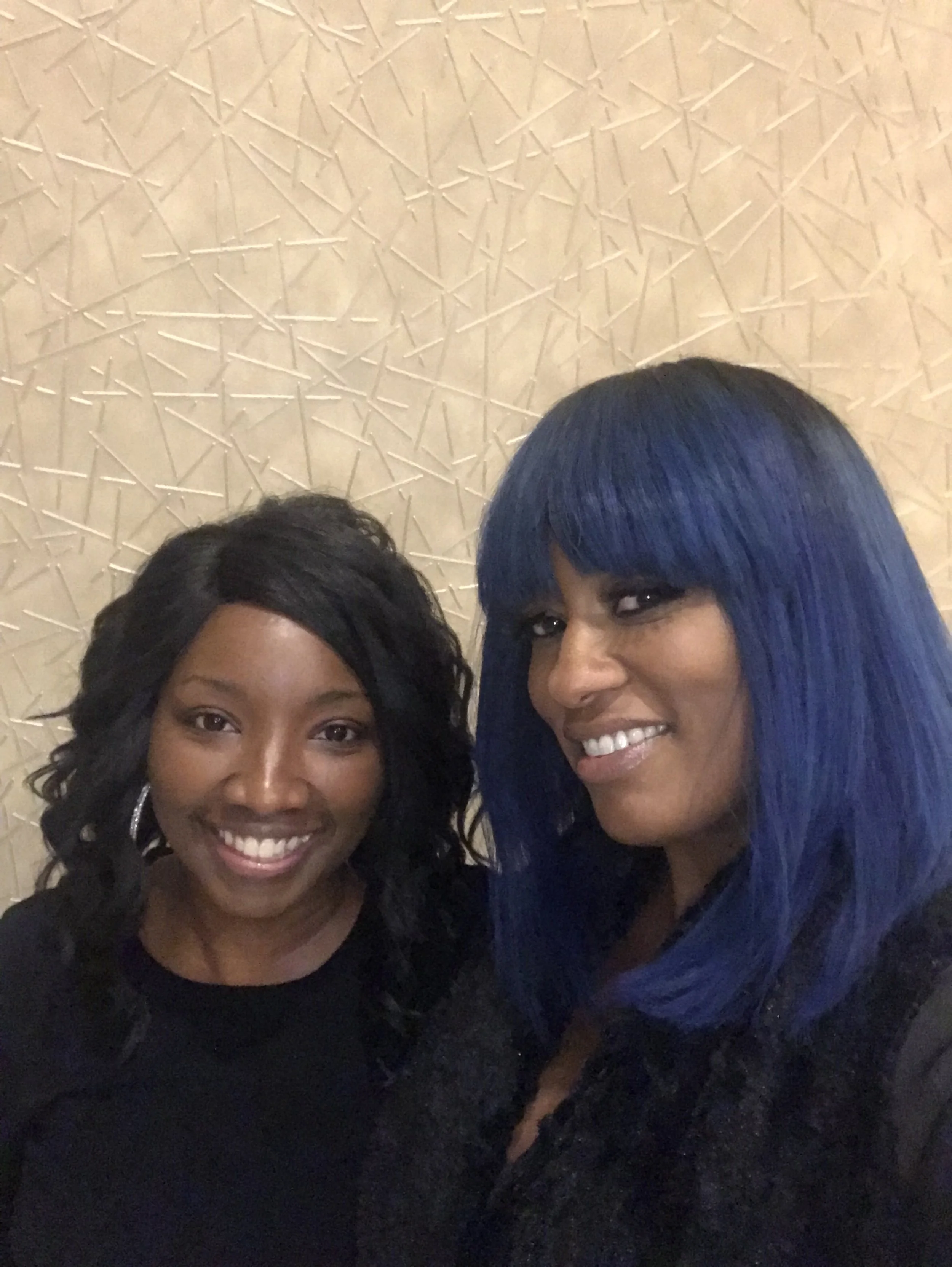 Two women are smiling and posing for a photo in front of a textured beige wall. The woman on the left has curly black hair and is wearing hoop earrings and a black top. The woman on the right has long, straight blue hair with bangs and is wearing a d