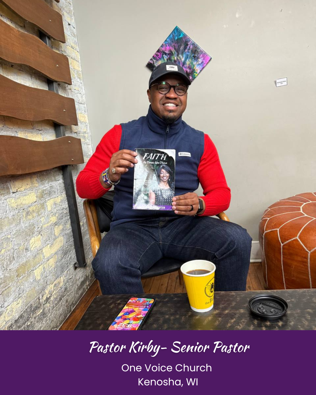 A man sitting indoors holding a book titled 'FAITH' at One Voice Church in Kenosha, WI. He's wearing glasses, a black cap, and a jacket with red sleeves. There is a yellow coffee cup, a smartphone, and a black lid on the table in front of him. The background shows a brick wall, a wooden wall feature, and abstract art hanging on the wall.