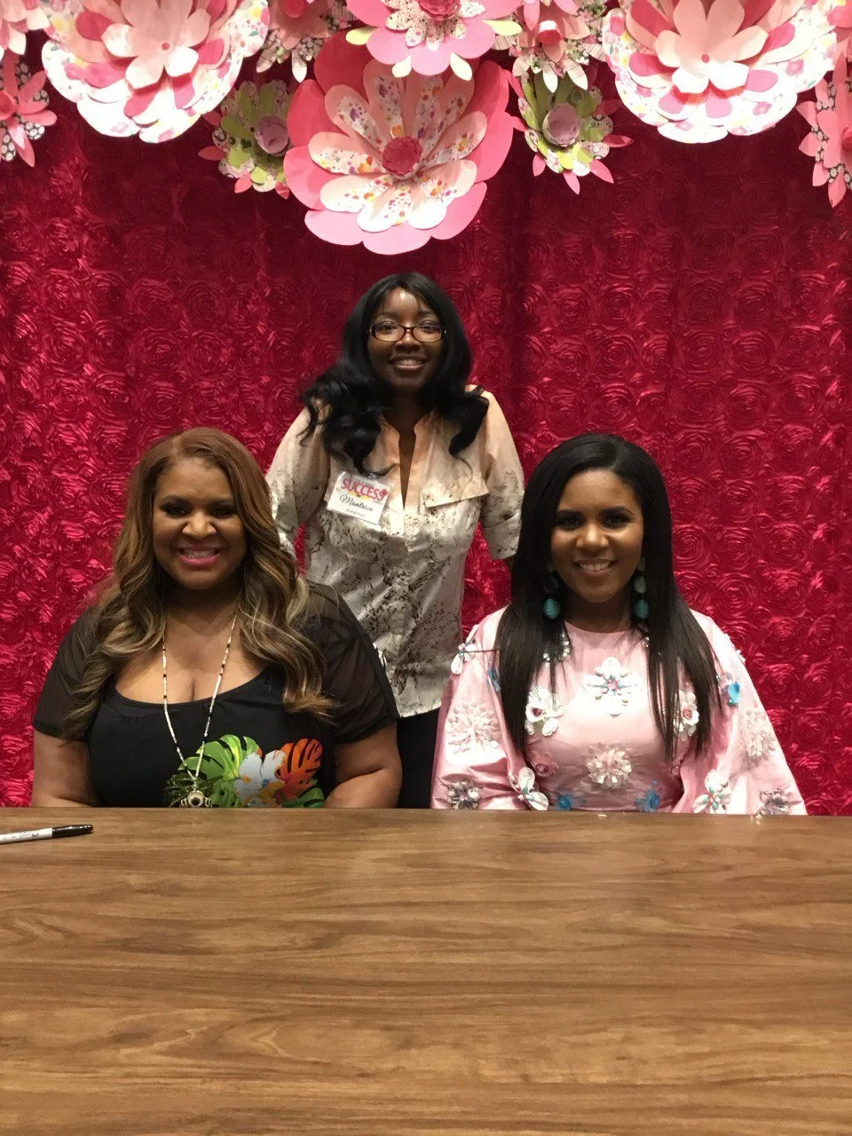 Three women sitting at a wooden table with a red textured backdrop and pink floral decorations overhead.