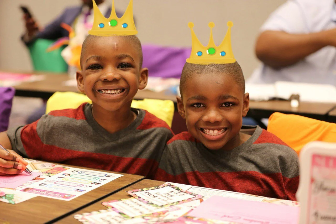 Two young boys with shaved heads are smiling, wearing red and gray shirts, each with a yellow paper crown decorated with green and blue circles. They are sitting at a table with colorful papers and school supplies, celebrating a special occasion in a