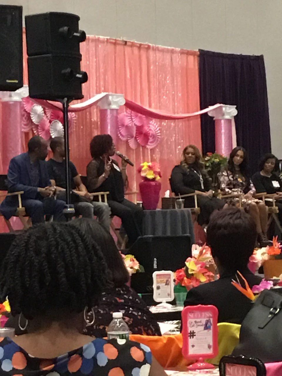 Panel of six women and men seated on stage during a conference or event, with a pink decorated backdrop, floral arrangements, and an audience in the foreground.