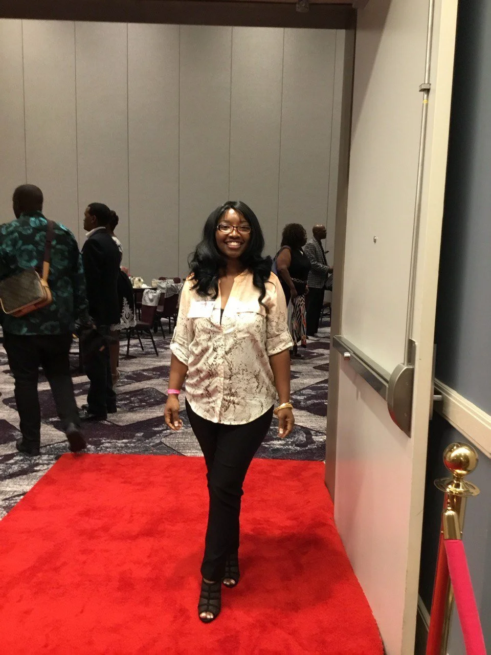 A woman walking on a red carpet at an indoor event, smiling and looking at the camera, with other people in the background.