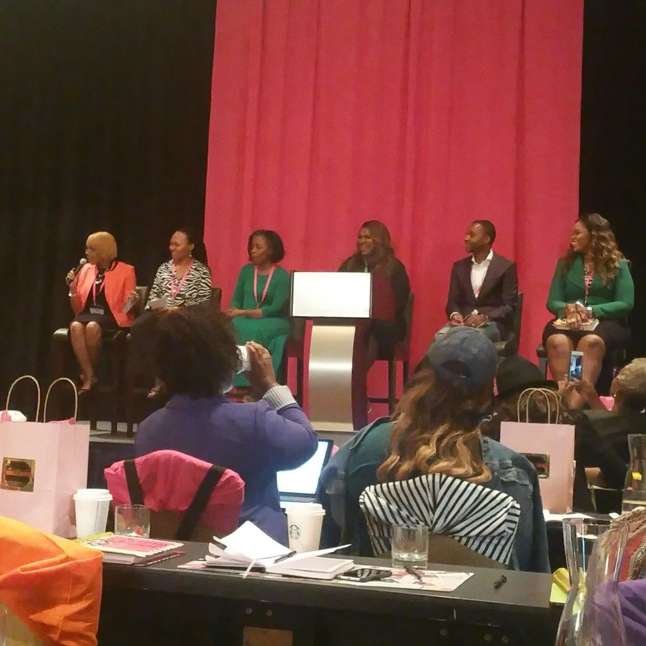 A panel discussion with six women and one man seated on stage in front of a pink backdrop, with one woman speaking into a microphone. Audience members are sitting at tables, some taking photos or using laptops.