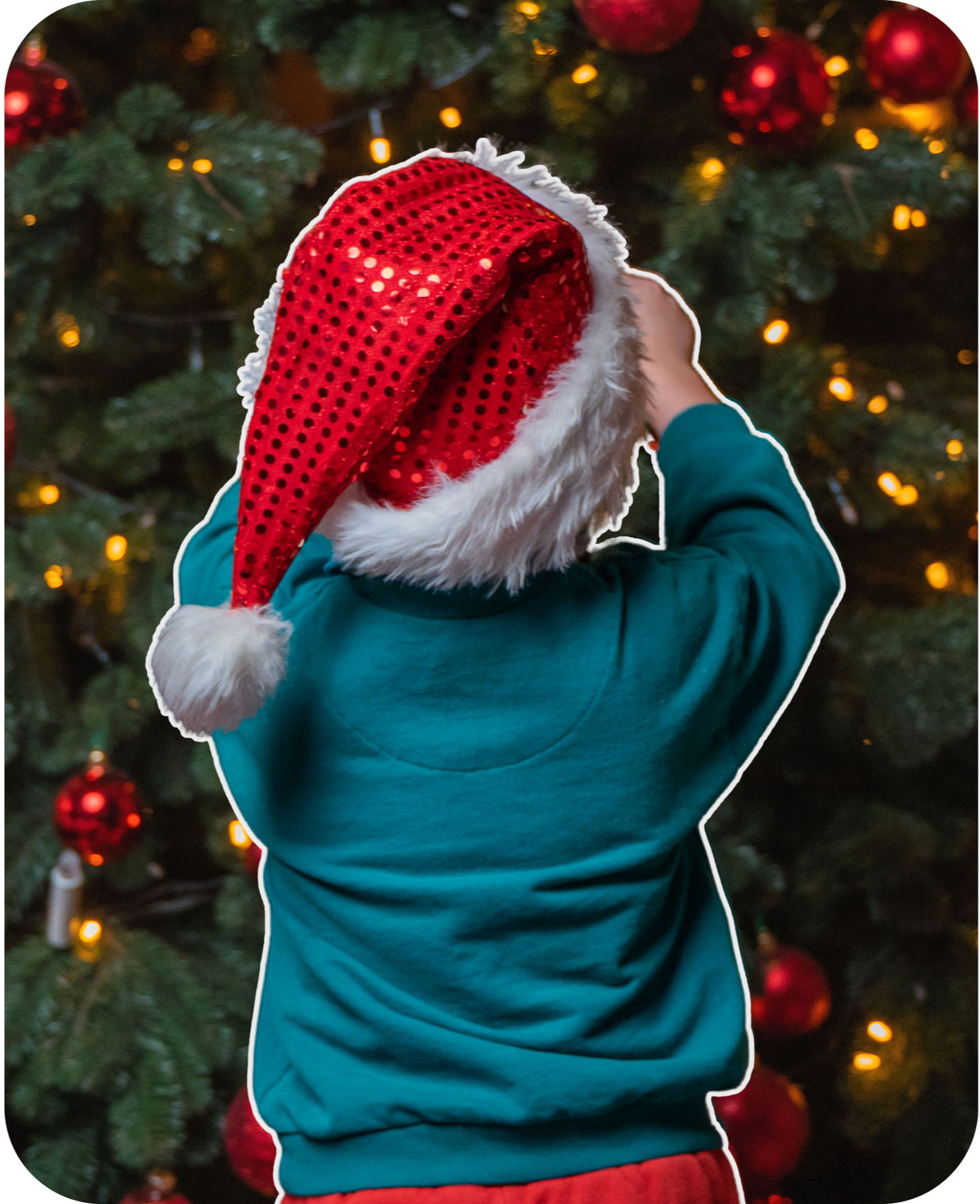 A child wearing a Santa hat and teal sweatshirt, standing in front of a decorated Christmas tree with red ornaments and yellow lights.